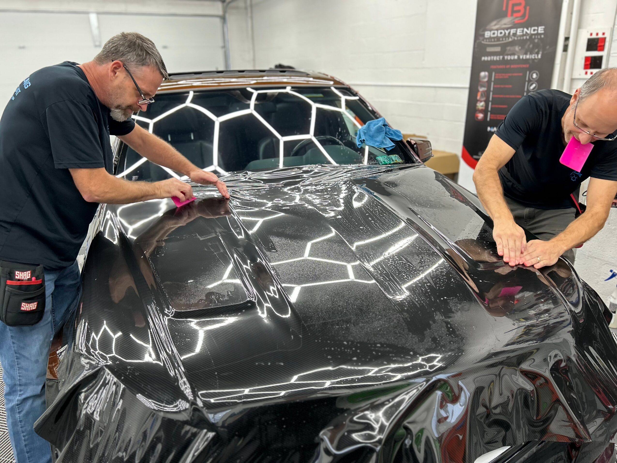 Two people are polishing a black car with a honeycomb pattern on the roof in a garage.