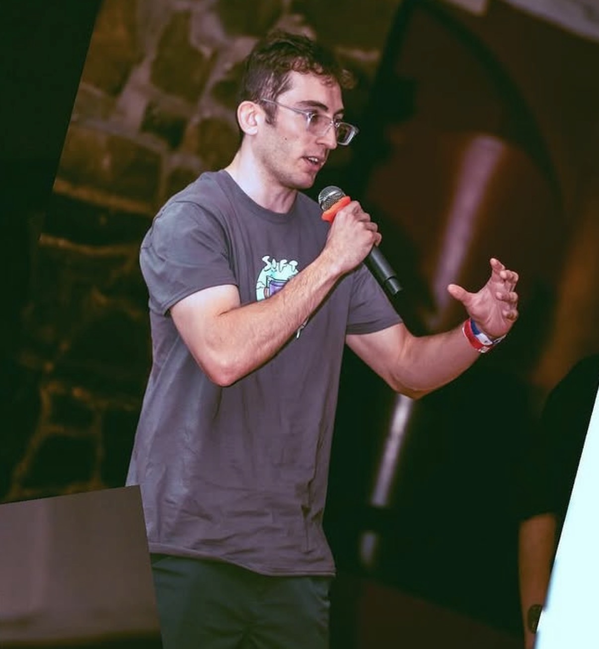 Young man with glasses speaking into a microphone, gesturing with his left hand, wearing a gray T-shirt, in a dimly lit setting.