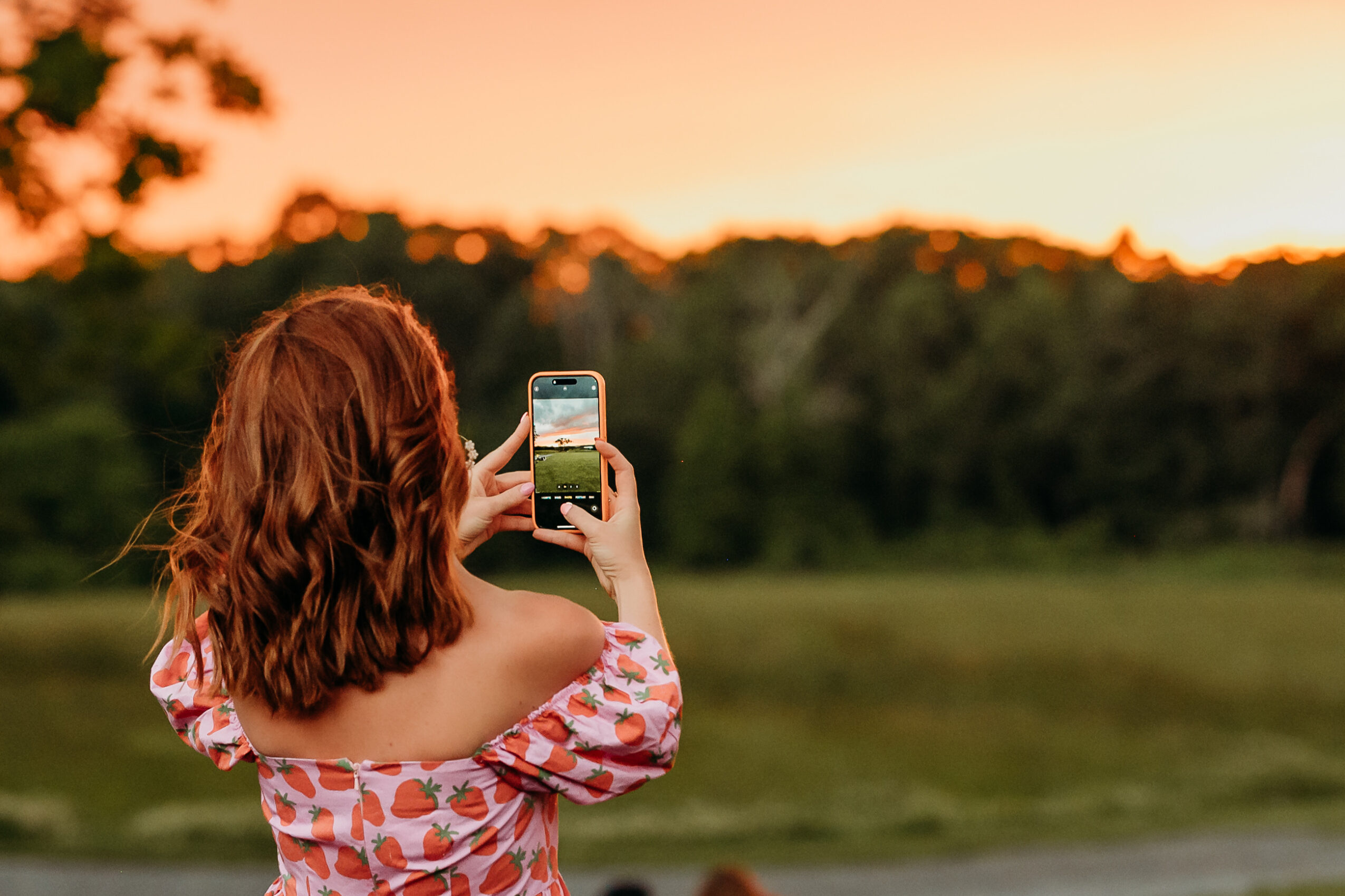 Person with shoulder-length hair taking a photo with a smartphone outdoors at sunset.