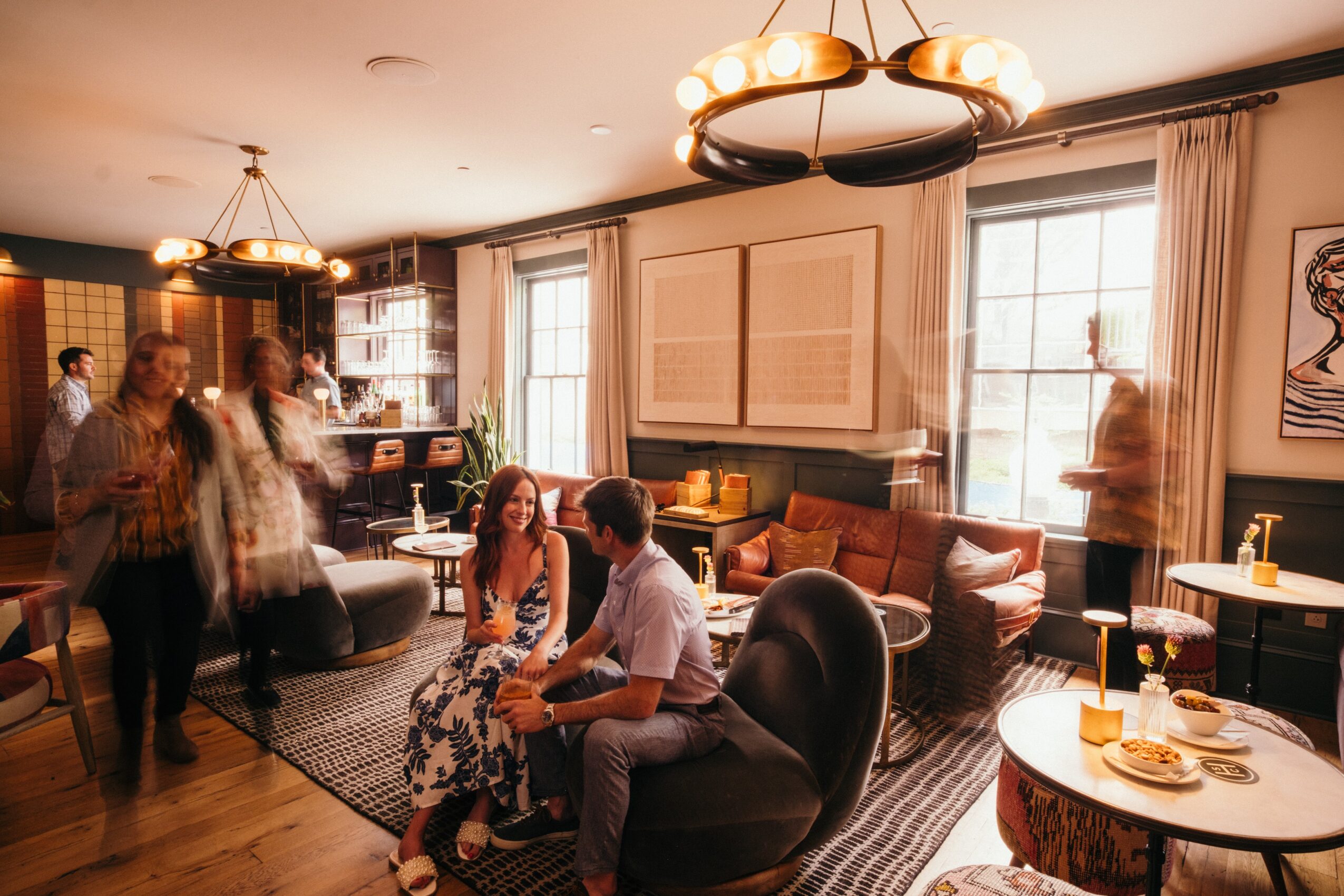 People socializing in a warmly lit living room with large windows, sofas, and a round table with food and drinks.