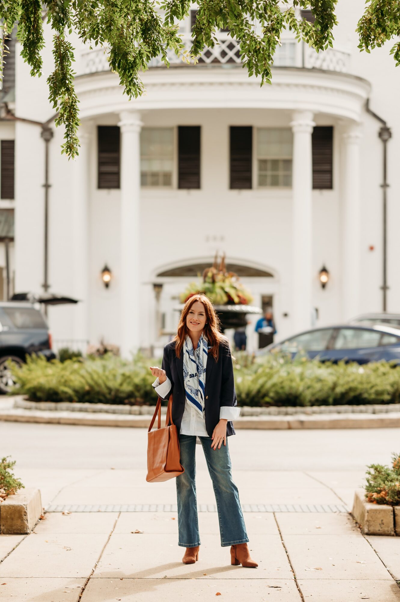 Woman standing on sidewalk in front of a large white building with columns, trees overhead, and parked cars nearby.