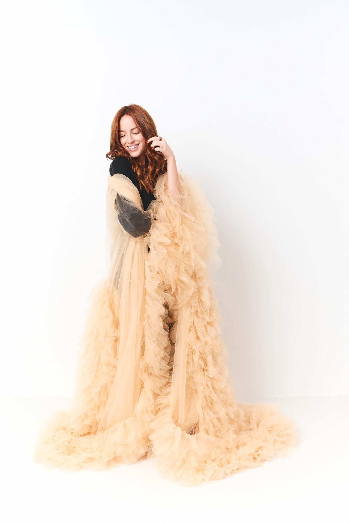 Woman with long hair smiling, wearing a beige fluffy gown, standing against a white background.