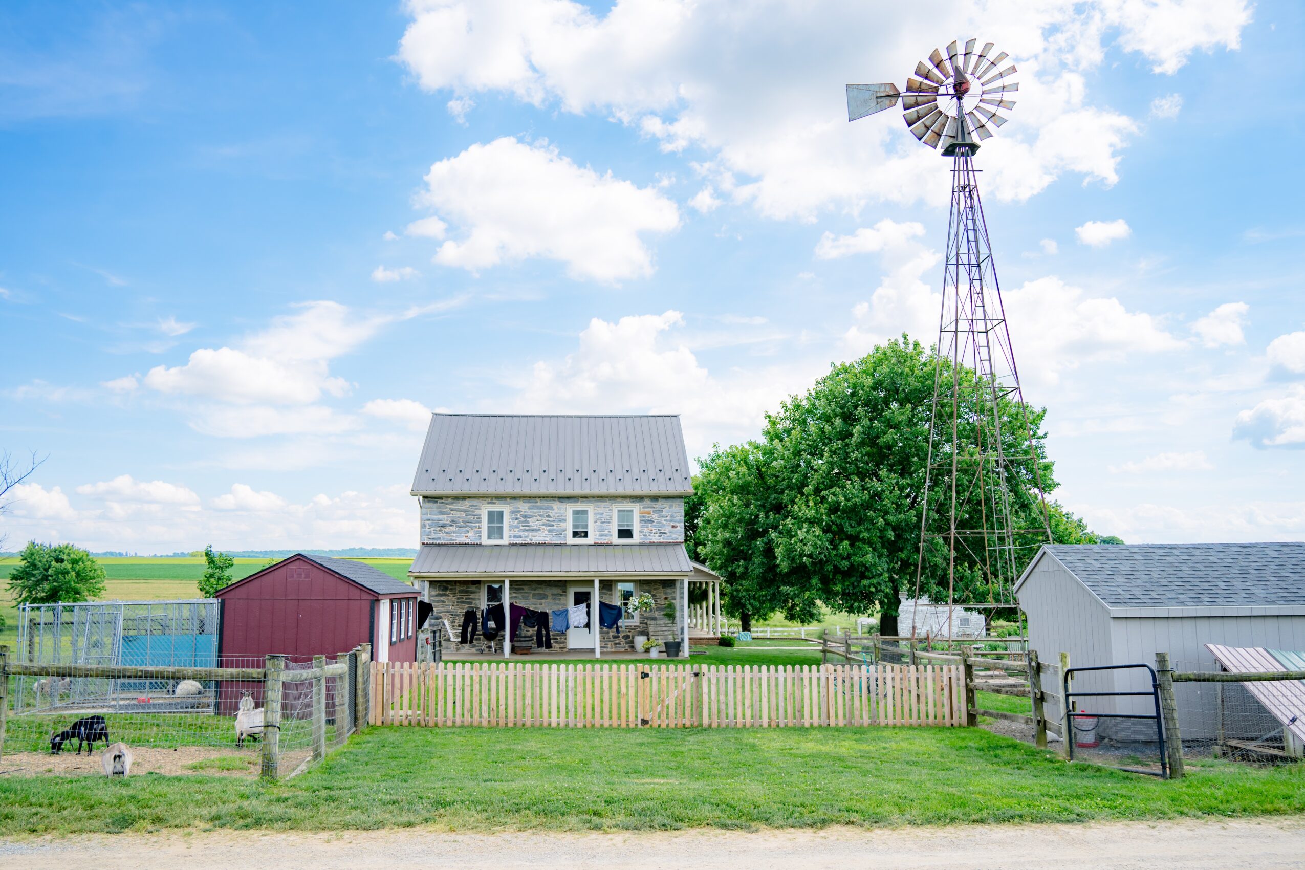 Rural scene with a house, windmill, trees, and small buildings under a partly cloudy sky.