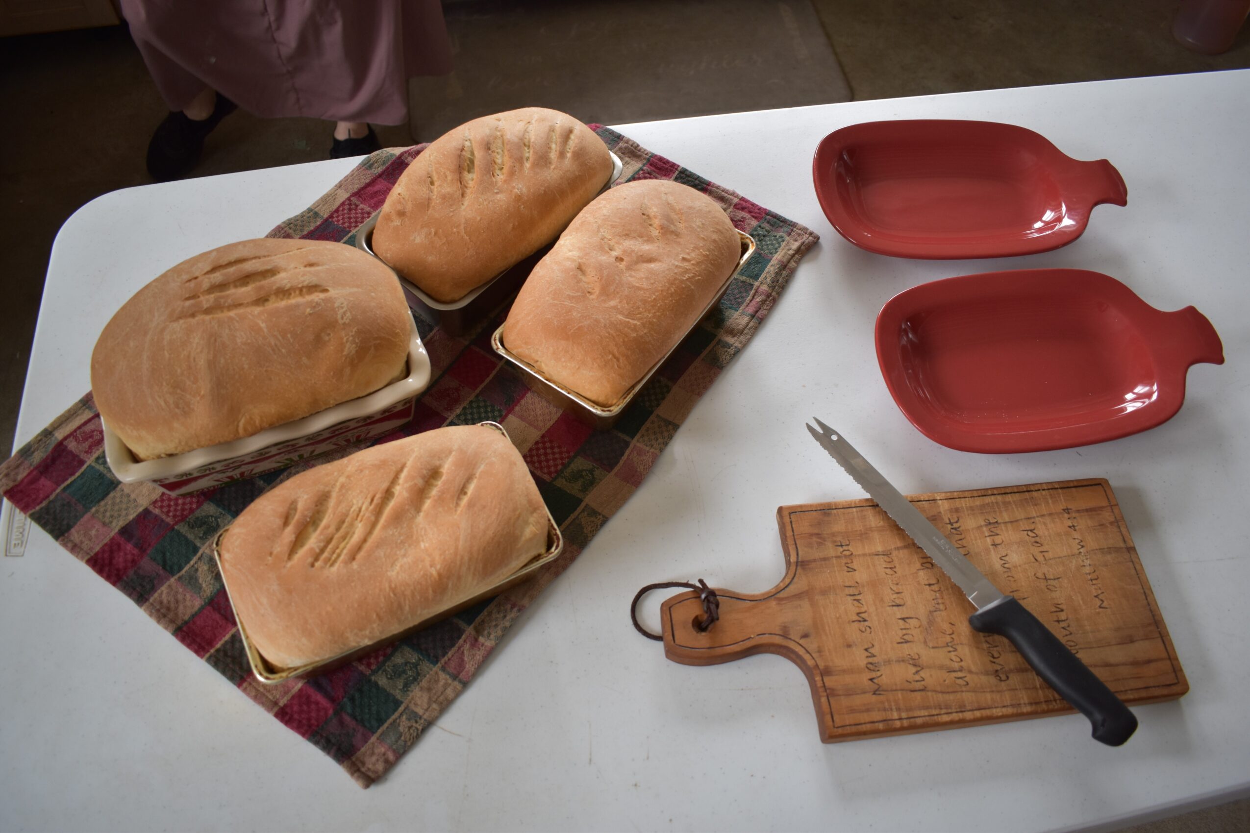 Four bread rolls on a checkered cloth, two red fish-shaped dishes, a wooden cutting board, and a knife on a white table.