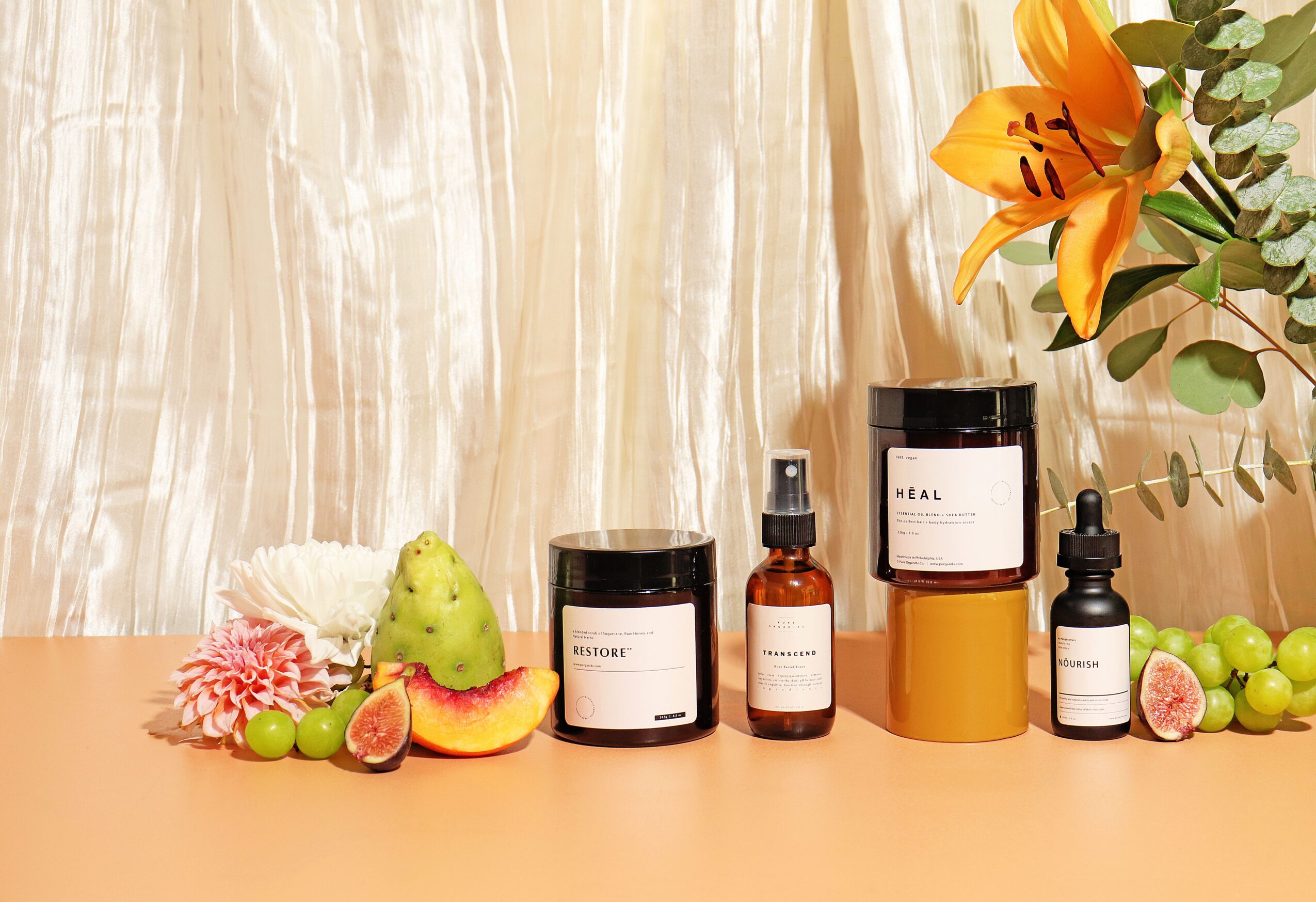 Arrangement of skincare products, fruits, and flowers on a surface with a curtain background.