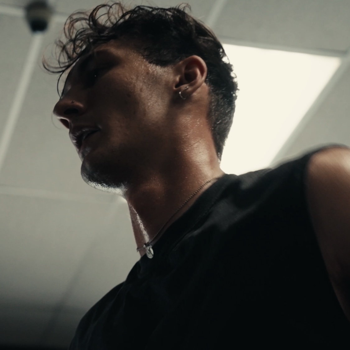 Side profile of a person with curly hair and a beard, wearing a dark shirt, in an indoor setting with ceiling lights.