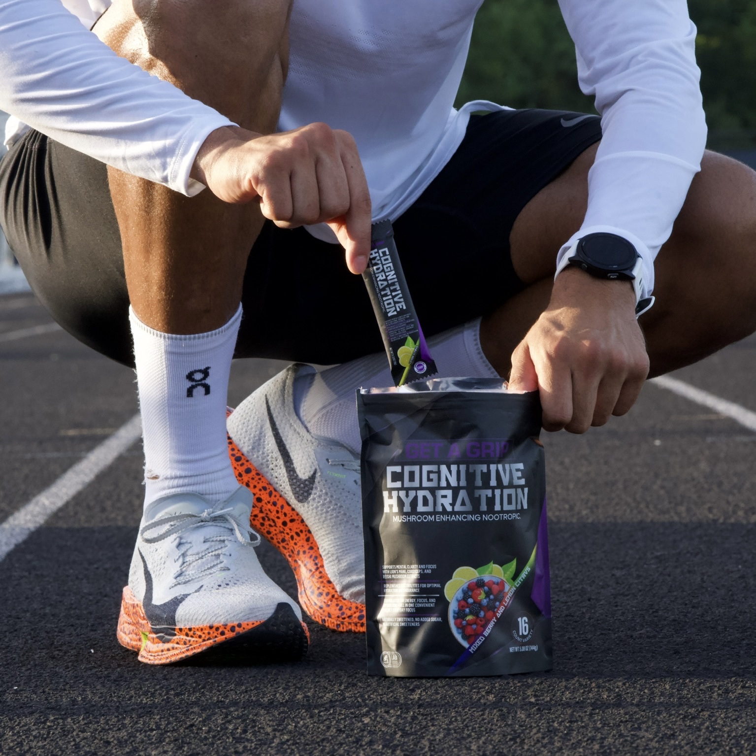 Person kneeling on track, opening a packet labeled 'Cognitive Hydration' with a purple and black design, outdoors.