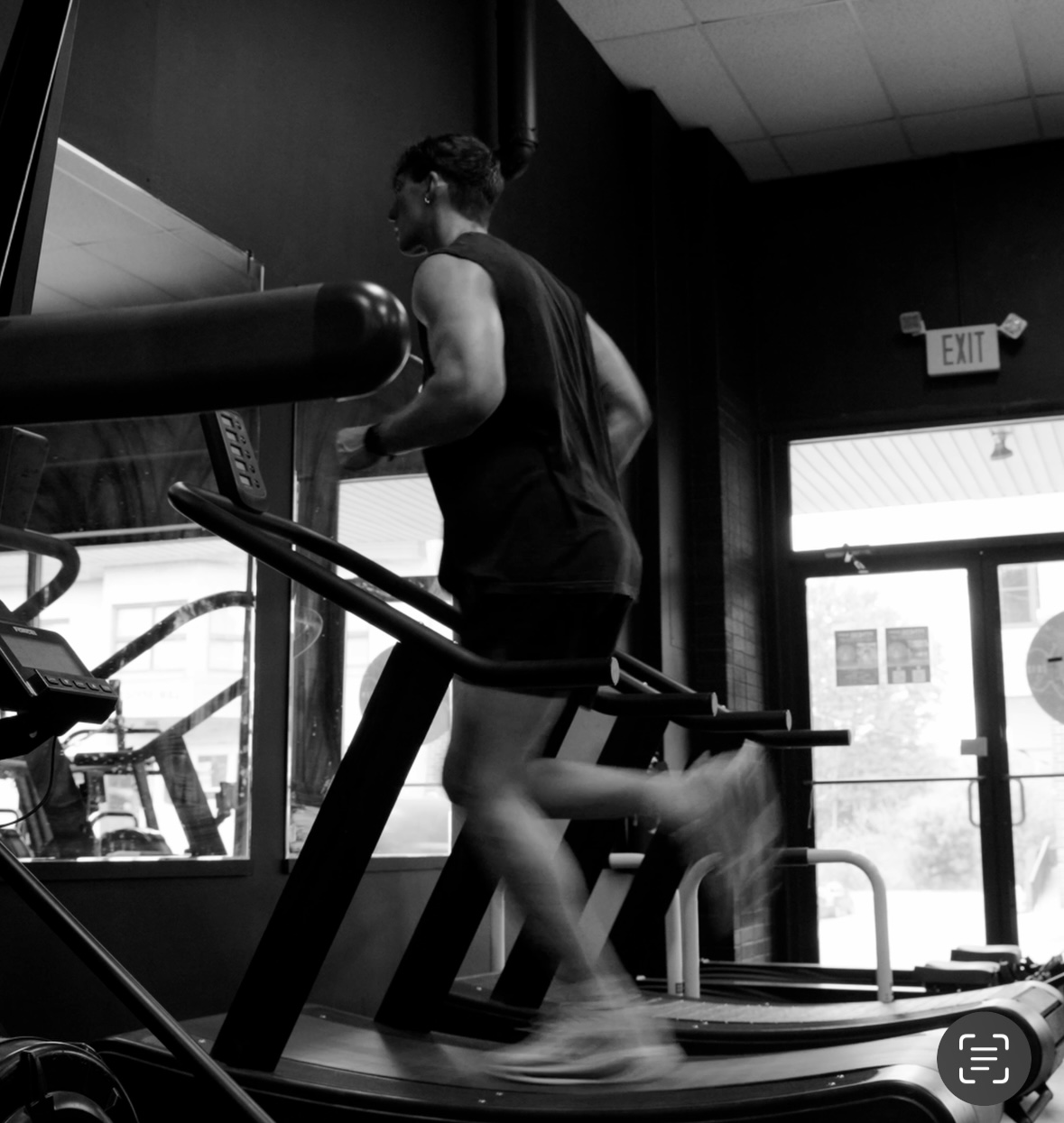 Person running on treadmill in gym, facing window, with exit door visible in background.