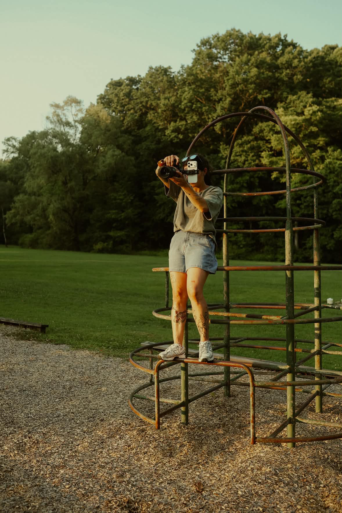 Person standing on playground equipment, holding a camera, with trees and grass in the background.