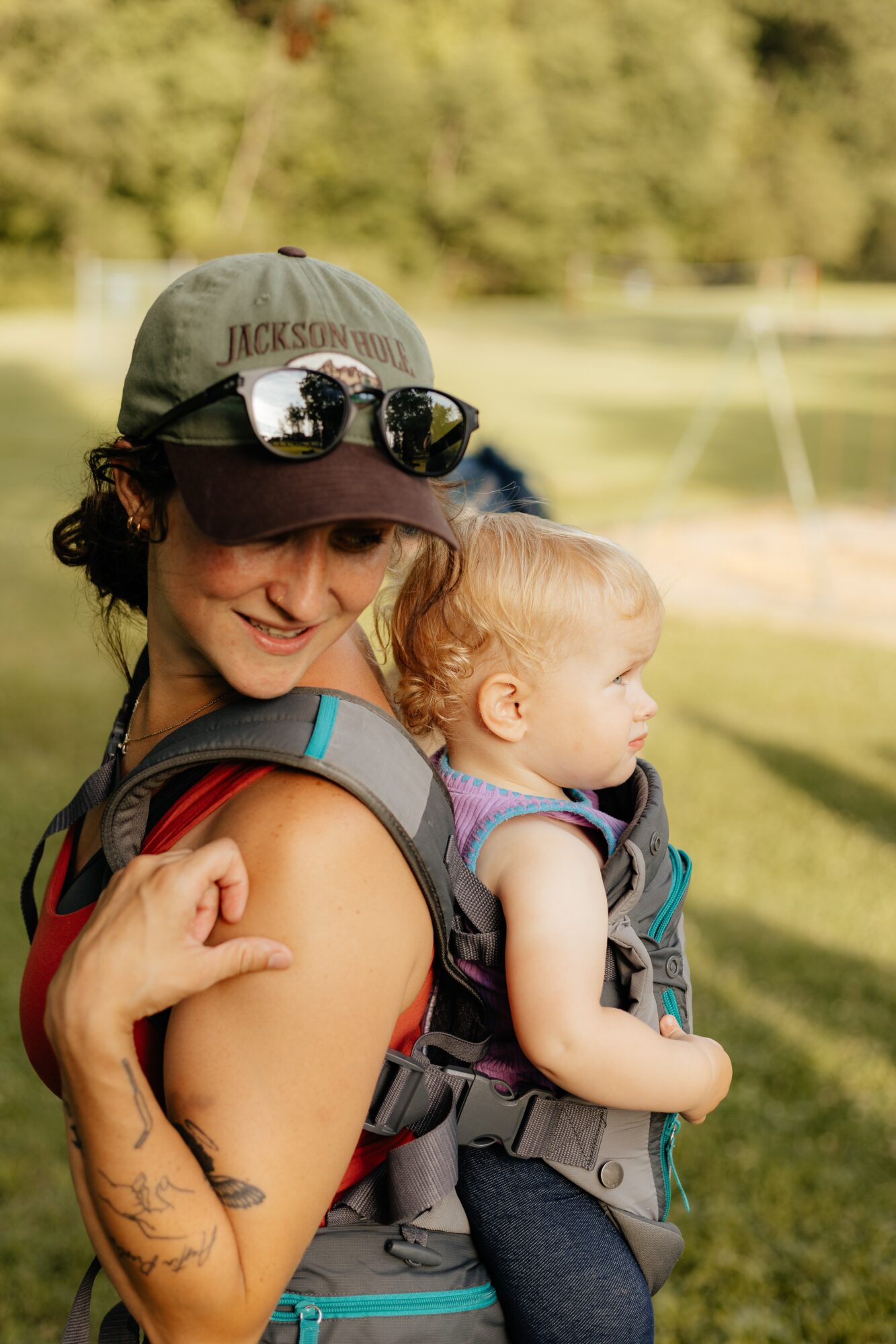 Woman with a child in a backpack carrier outdoors, smiling, with trees and grass in the background.