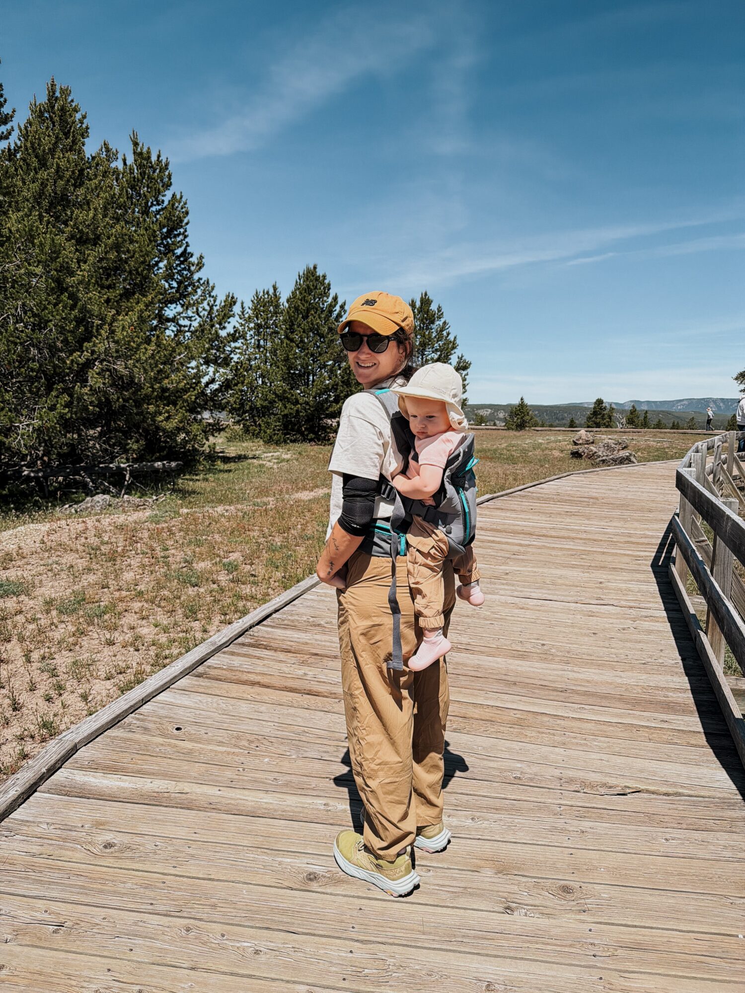 Person carrying a child on a wooden bridge outdoors with trees and blue sky in background.