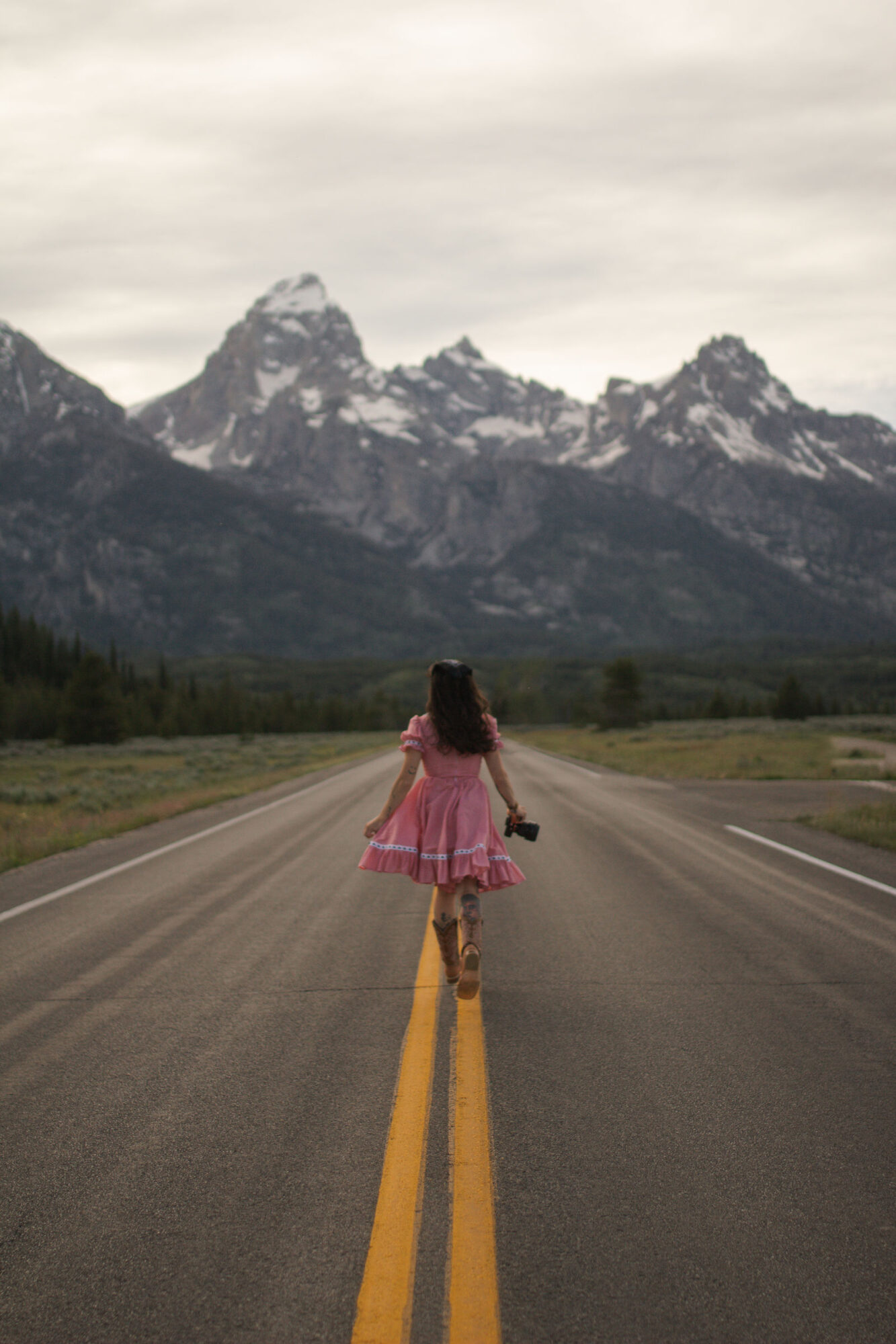 Person walking on a road toward mountains, wearing a pink dress and holding a camera, with a cloudy sky.