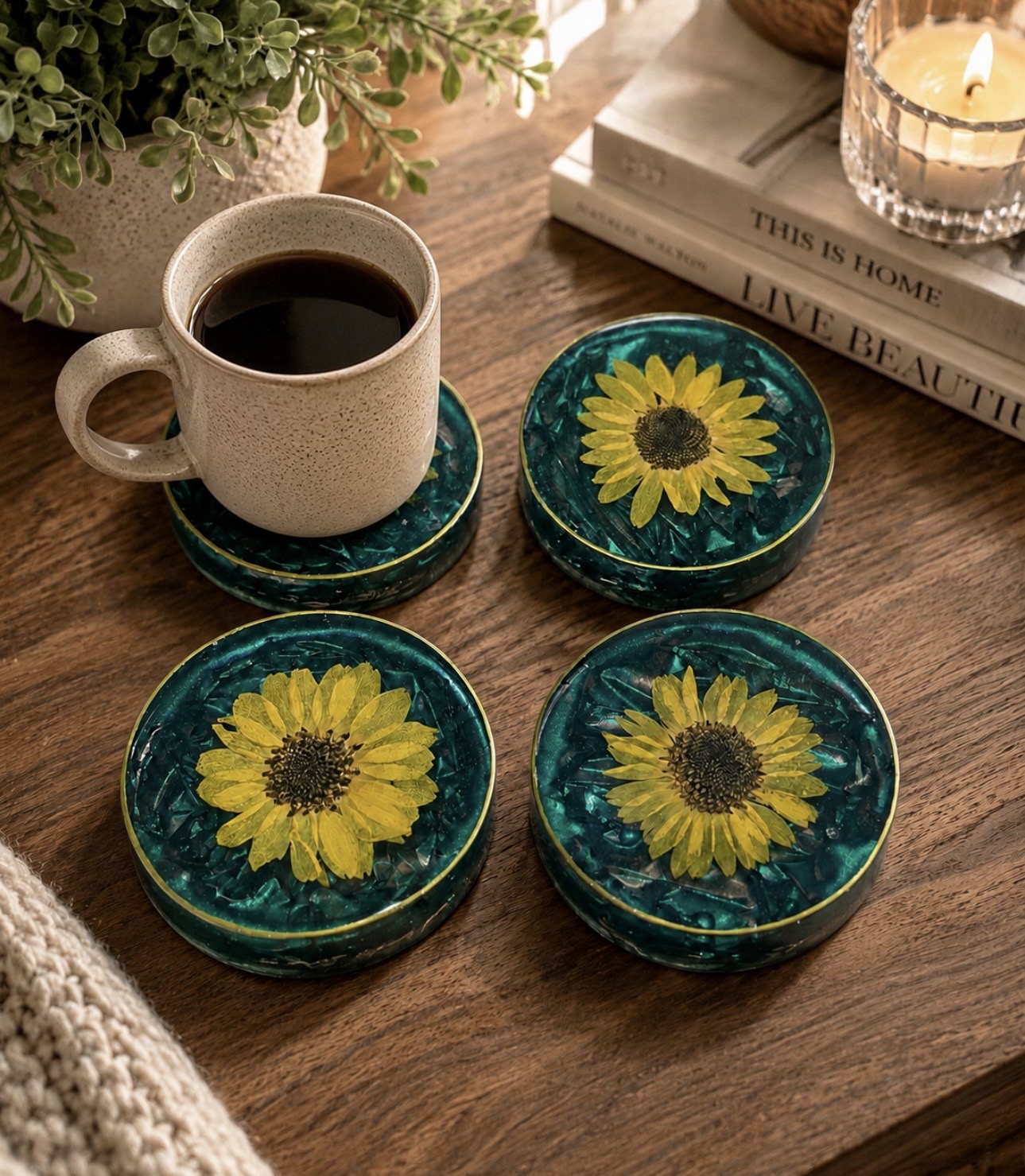 Coffee mug on a coaster with sunflower design, four sunflower coasters, books, and a lit candle on a wooden table.