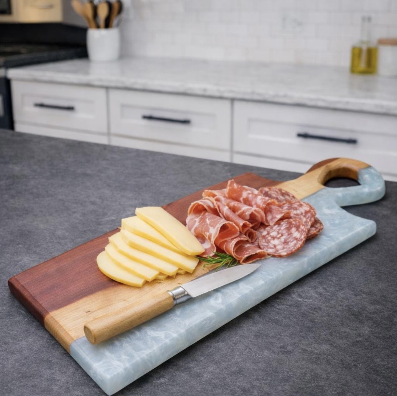 Cheese and cured meats on a wooden and marble serving board in a kitchen, with a gray countertop and white cabinets in the background.