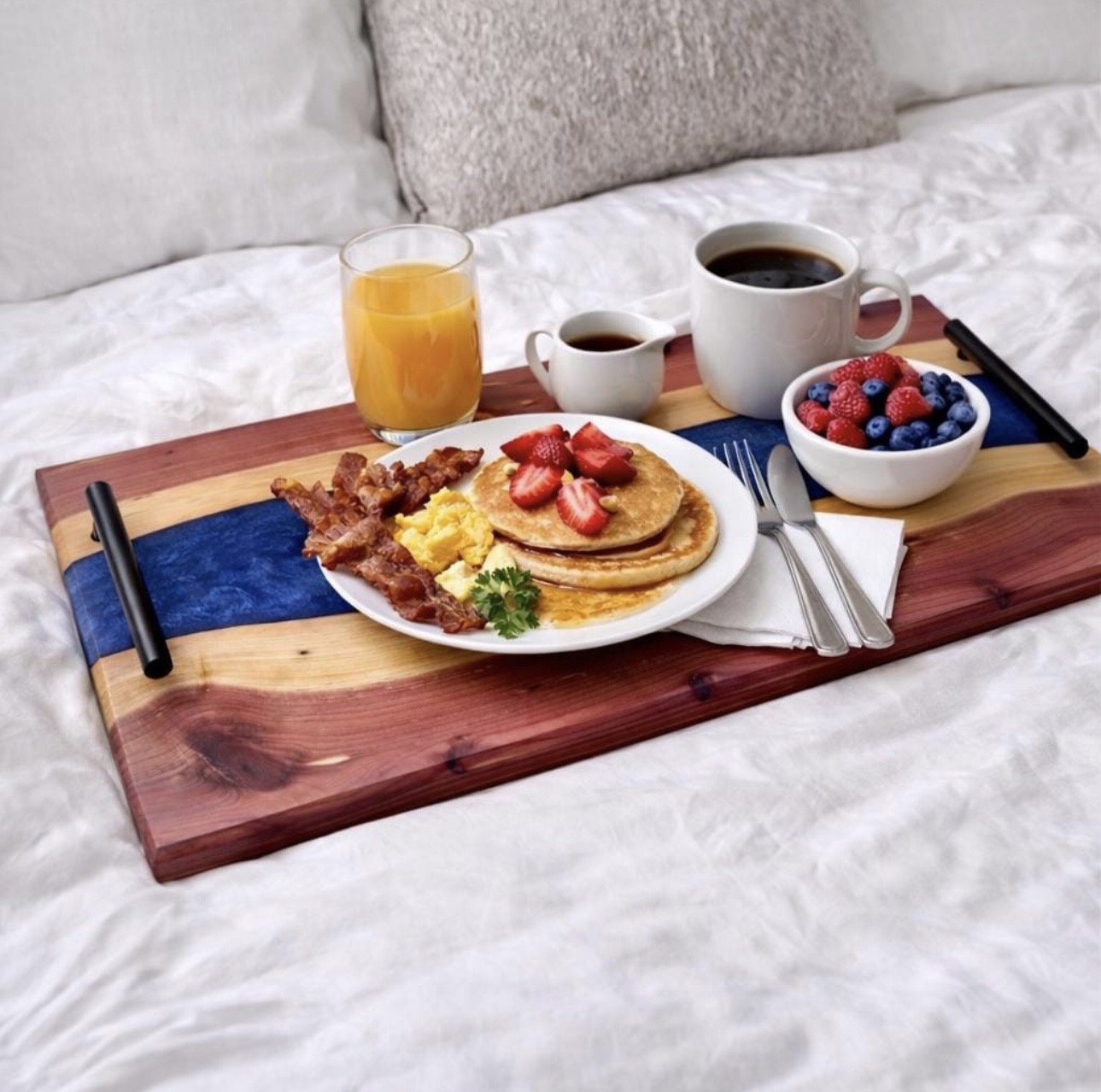 Breakfast tray with pancakes, strawberries, bacon, scrambled eggs, orange juice, coffee, and berries on a bed.