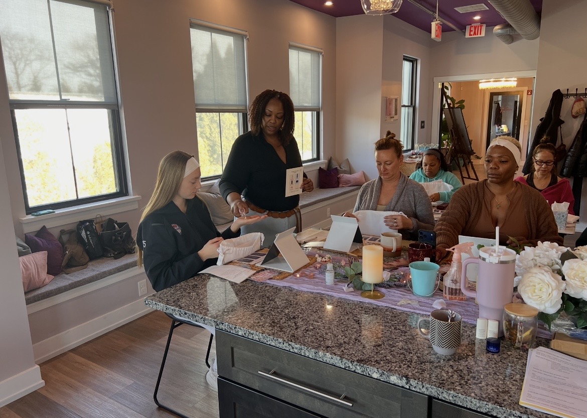 Group of women sitting around a kitchen island, reading papers, with a woman standing and speaking, in a bright room.