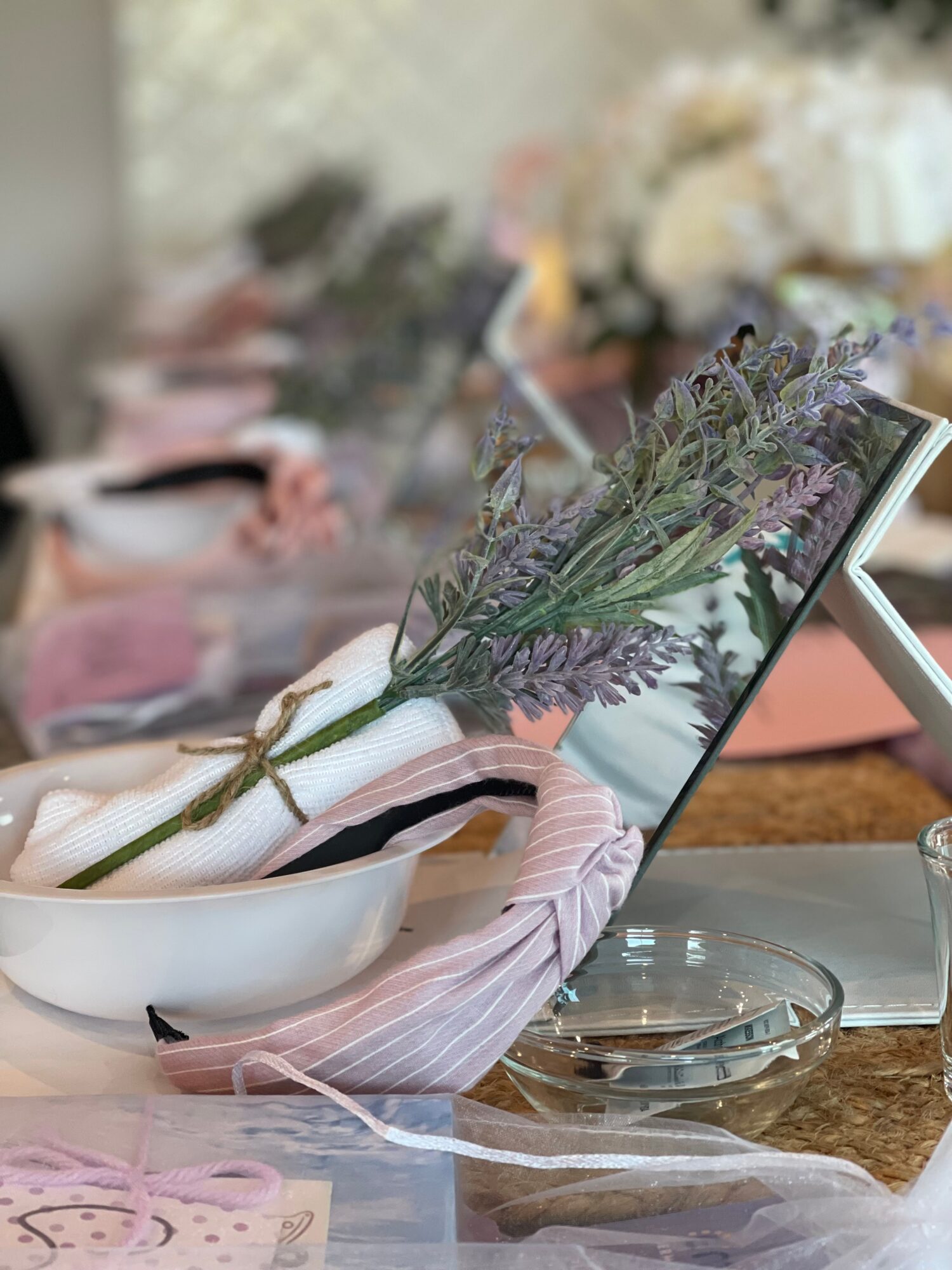 A bouquet of lavender and greenery in a white bowl, with a mirror reflecting the arrangement, on a table.