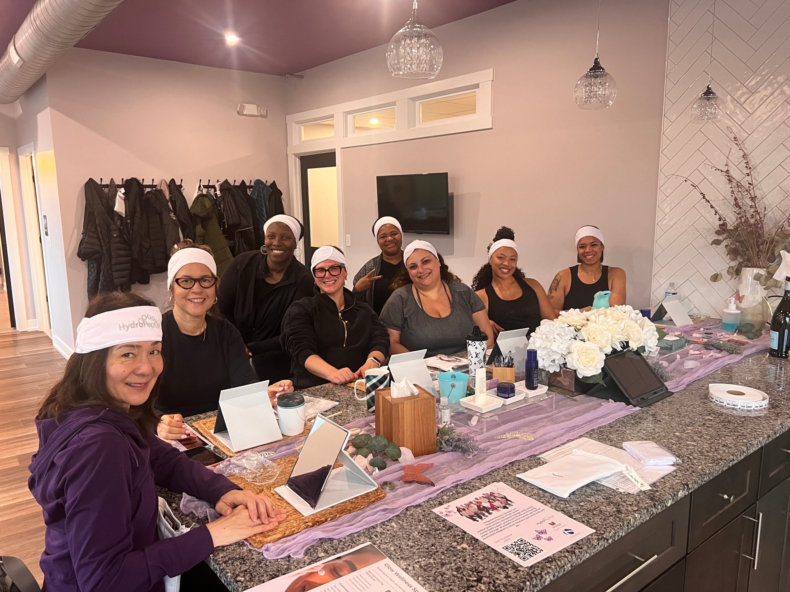 Seven women wearing white head coverings sit at a kitchen counter with flowers and papers, smiling at the camera.