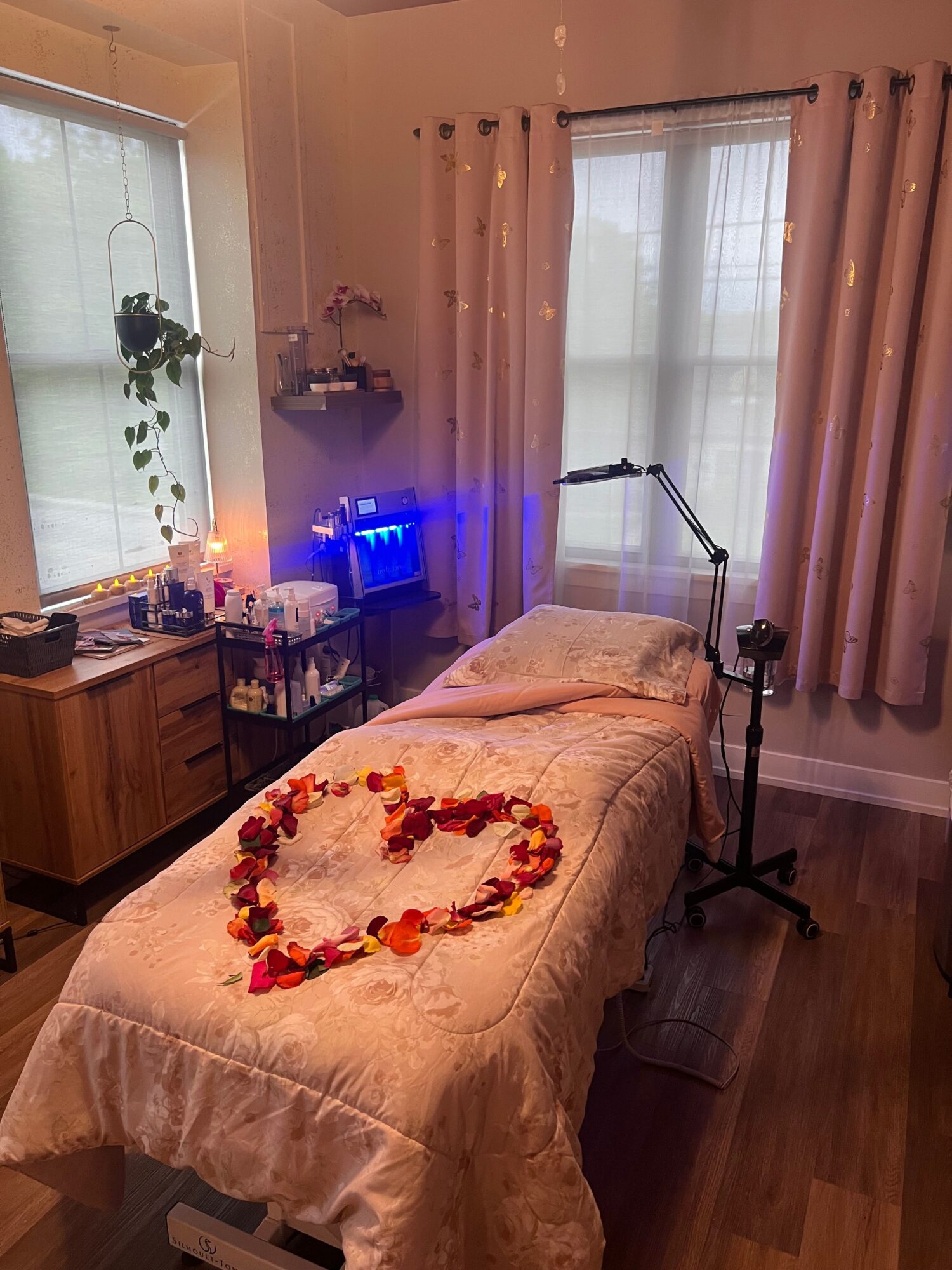 A room with a bed, a window with curtains, a desk with a computer, and a heart shape made of flowers on the bed.
