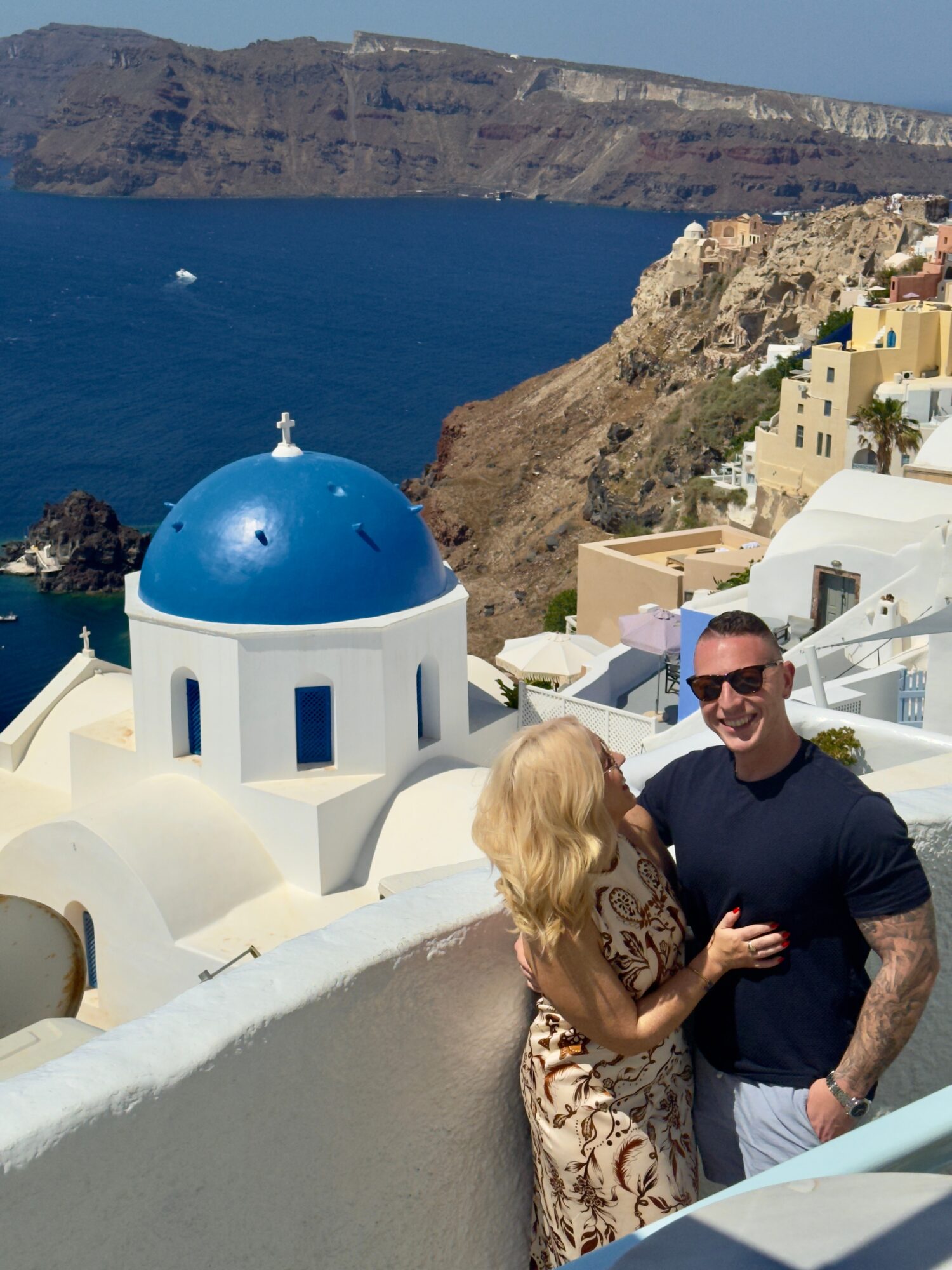 Two people, a man and a woman, stand close together on a white wall with a blue-domed building and coastal cliffs in the background.