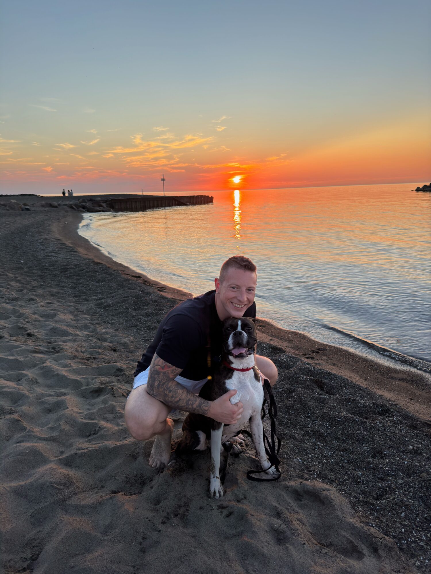 Person kneeling on beach holding a dog with sunset over water in background.