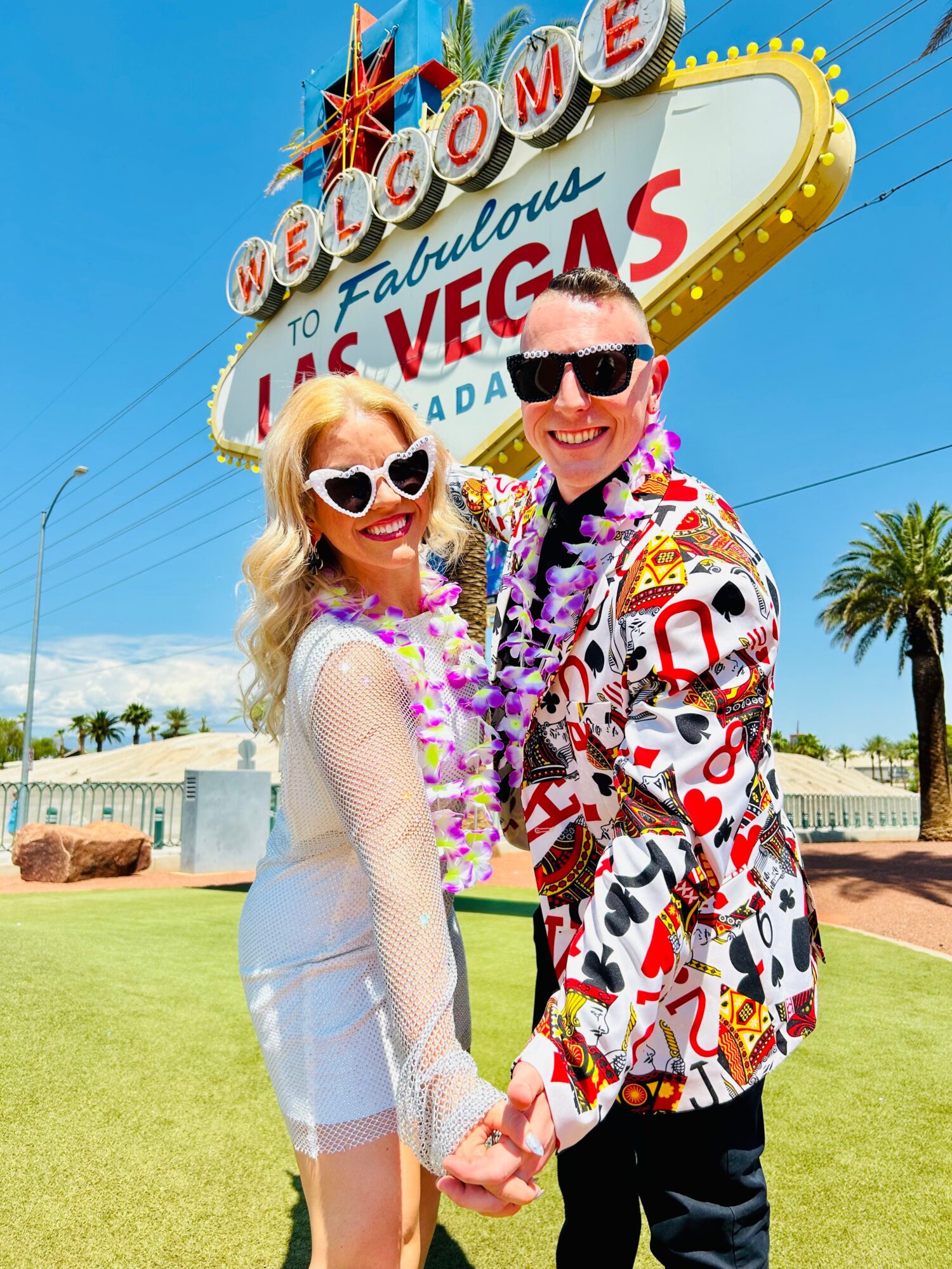 A smiling couple holding hands in front of the Las Vegas welcome sign, wearing sunglasses and colorful clothing.