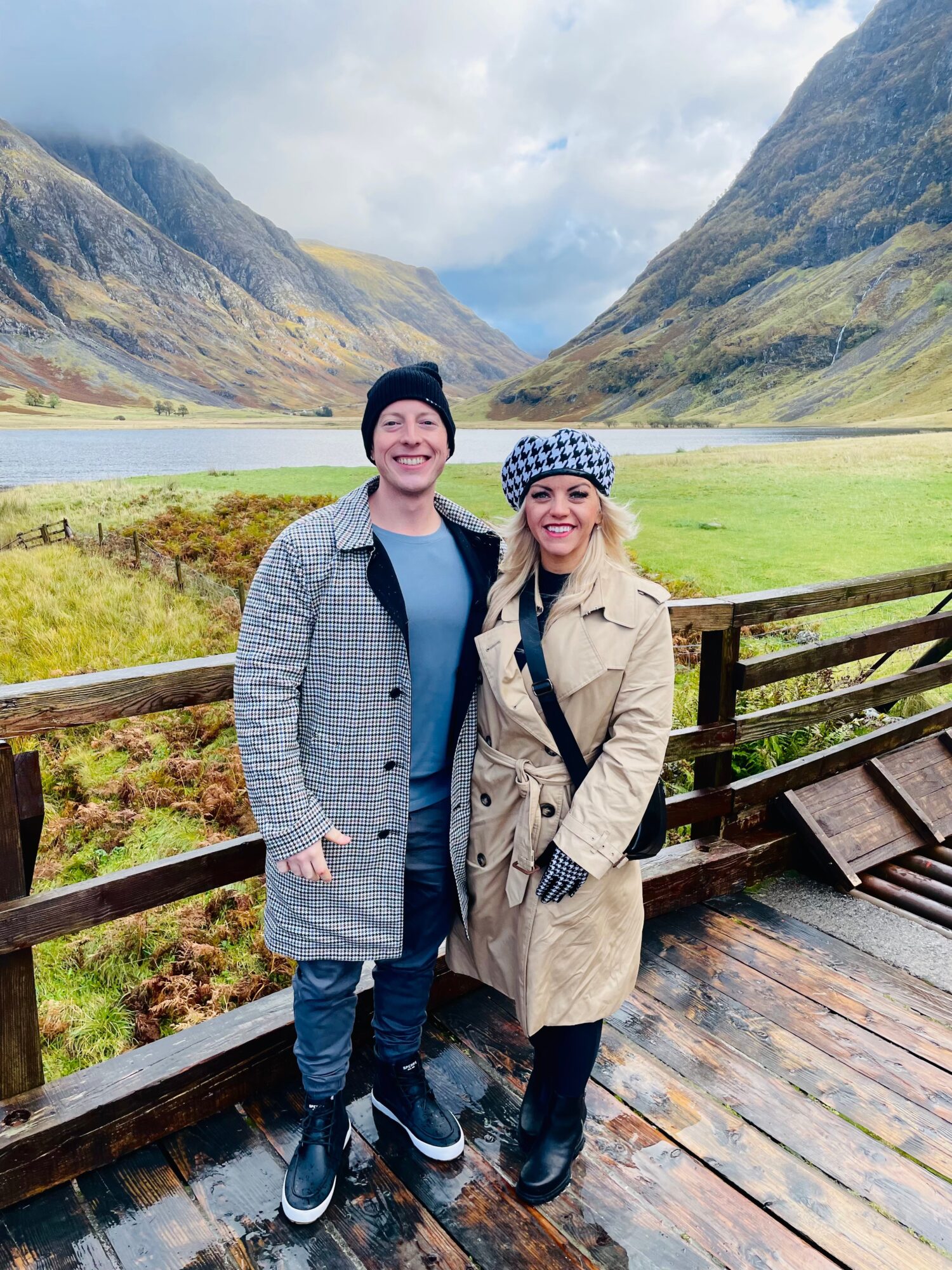 A smiling man and woman stand on a wooden platform with a scenic valley, mountains, and a lake in the background.
