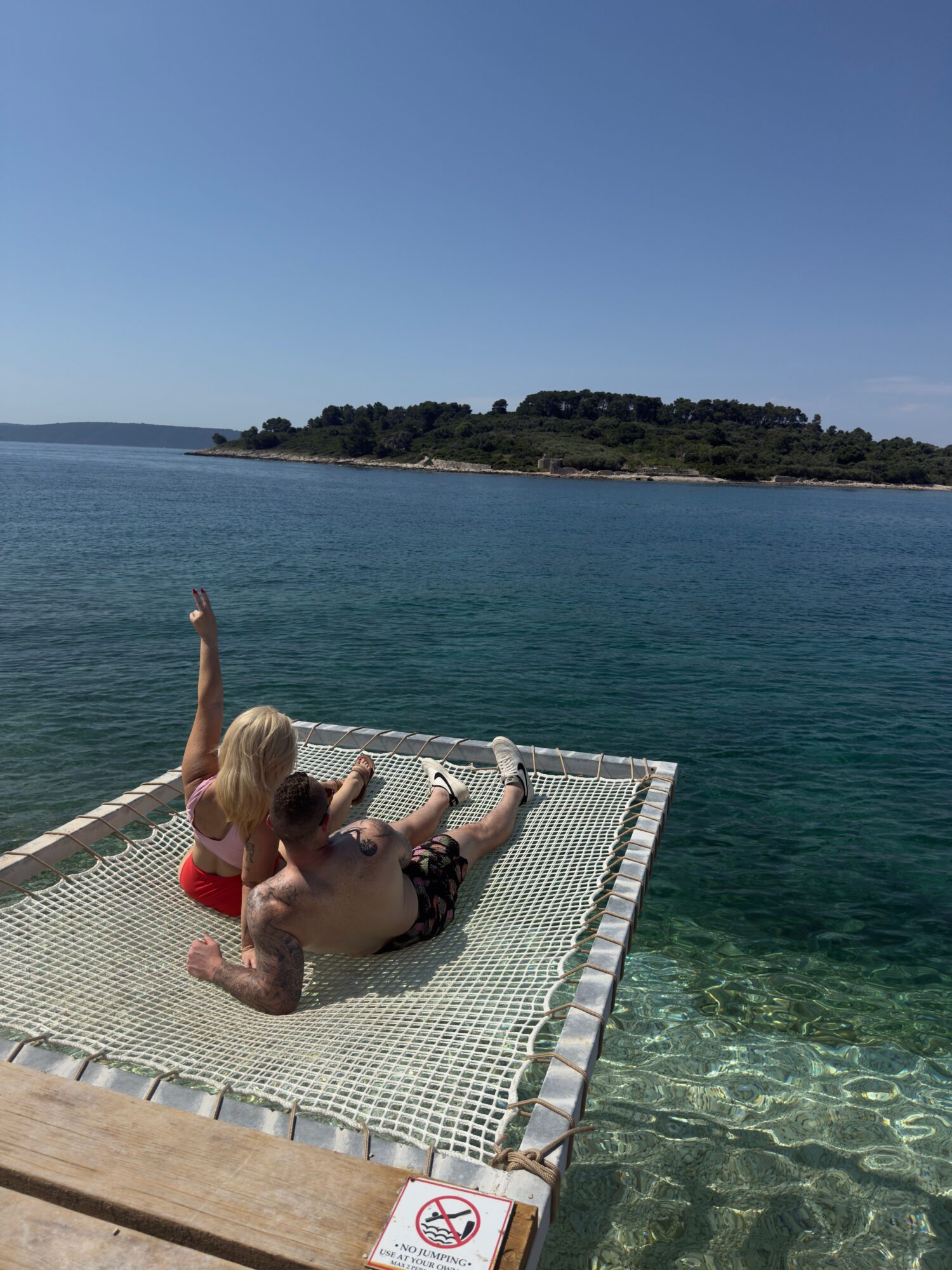 Two people relaxing on a floating dock by the water, with a person raising an arm, and a distant landmass in the background.