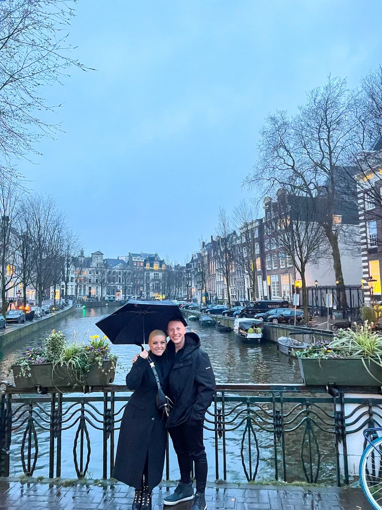 Two people stand by a canal with boats, buildings, and leafless trees in the background on a cloudy day.