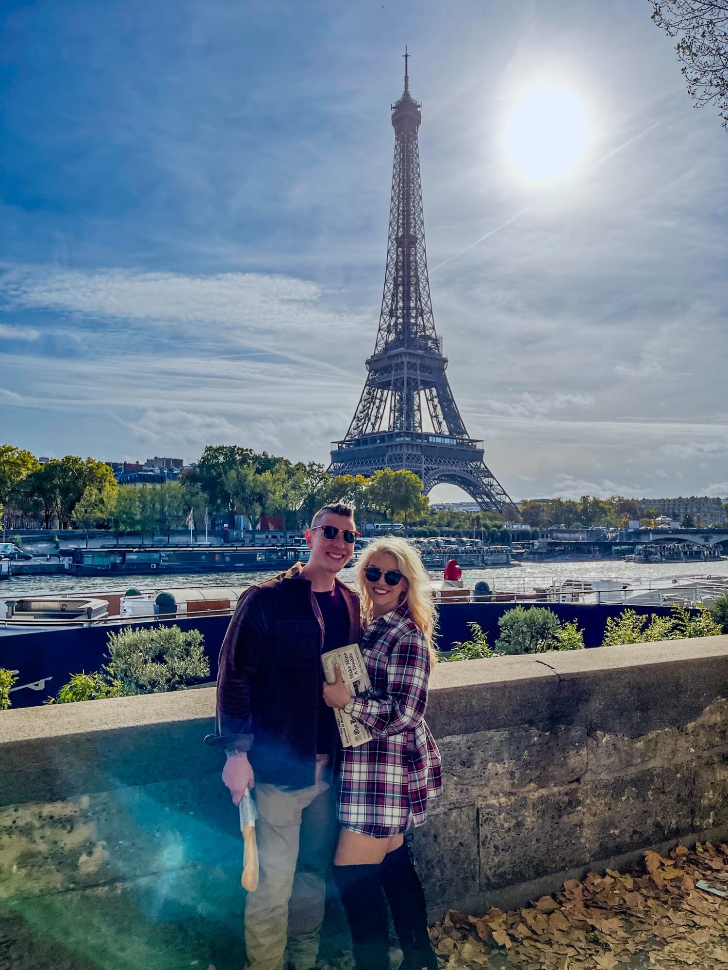 Two people stand in front of the Eiffel Tower with a river and boats behind them, sunny sky, and trees in the background.