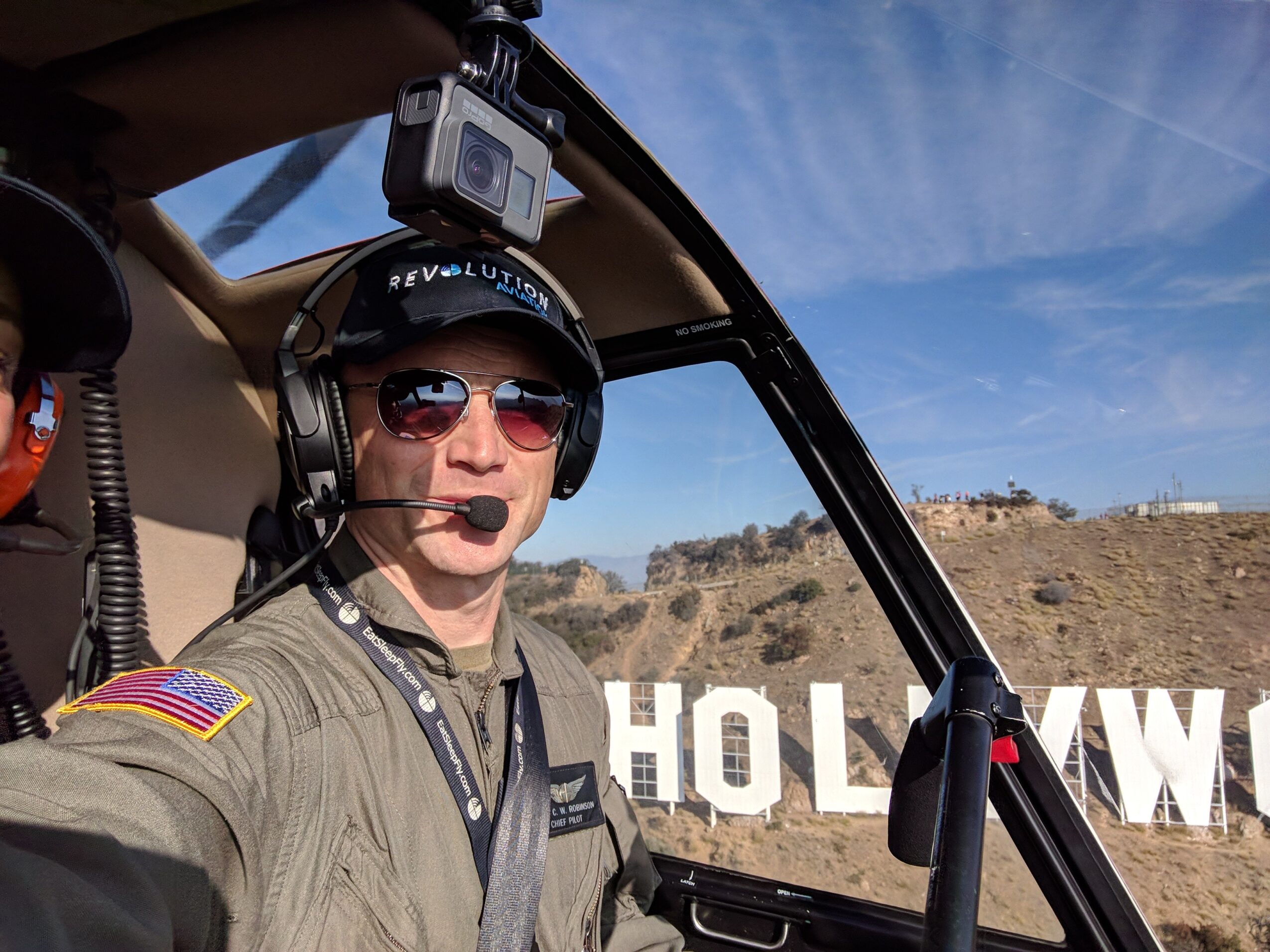 Person in pilot uniform with sunglasses and headset inside helicopter, Hollywood sign visible outside, clear blue sky.
