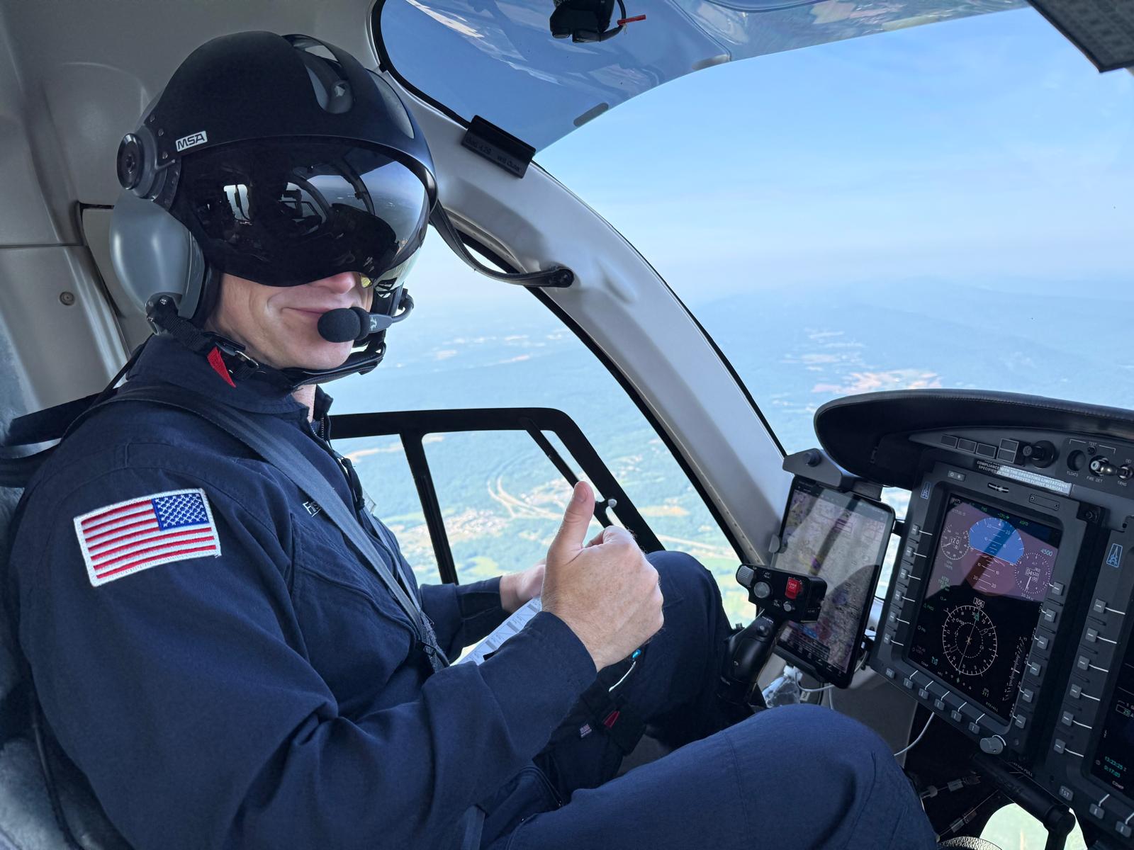 Pilot in cockpit wearing helmet and headset, giving thumbs up, with aircraft controls and sky visible outside window.