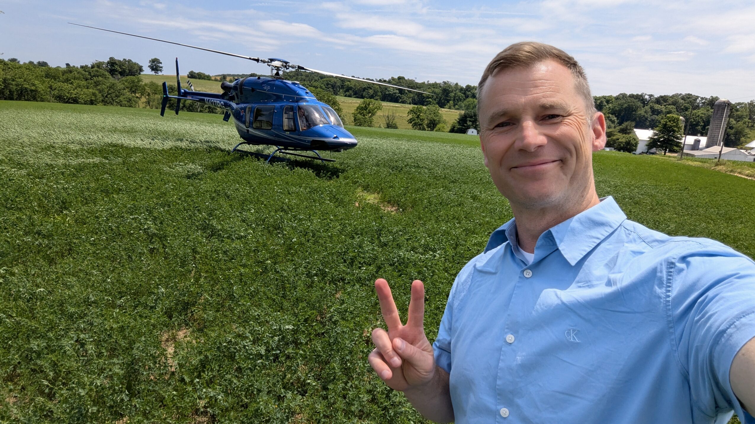 Man taking selfie outdoors with a blue helicopter in the background, holding up a peace sign, green field and trees behind.