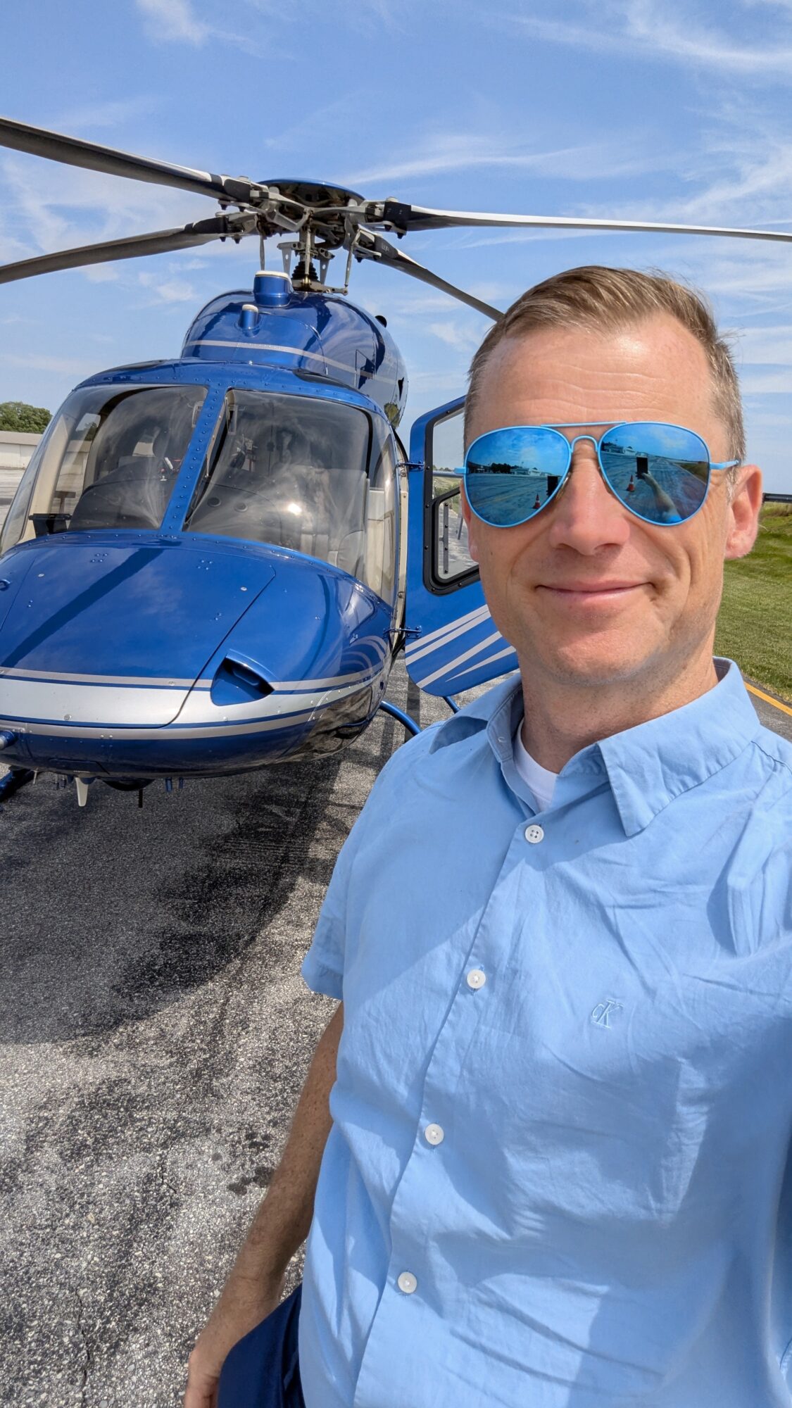 Man wearing sunglasses and a light blue shirt standing in front of a blue helicopter on a sunny day.