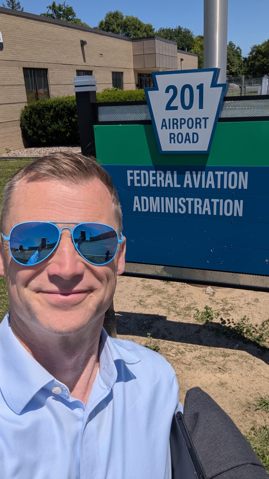 Person wearing sunglasses and a light blue shirt taking a selfie outdoors near a sign for Airport Road and Federal Aviation Administration.