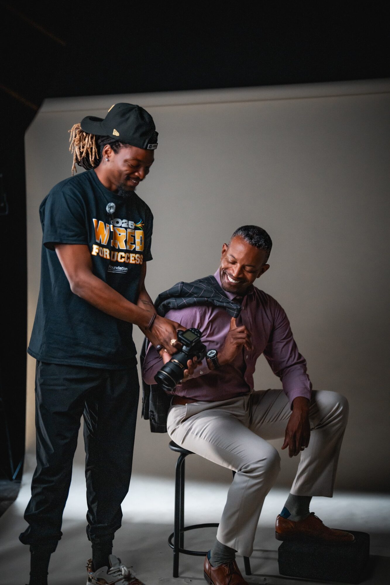 Two men interact; one holds a camera, the other sits on a stool, smiling, in a studio setting with a plain background.