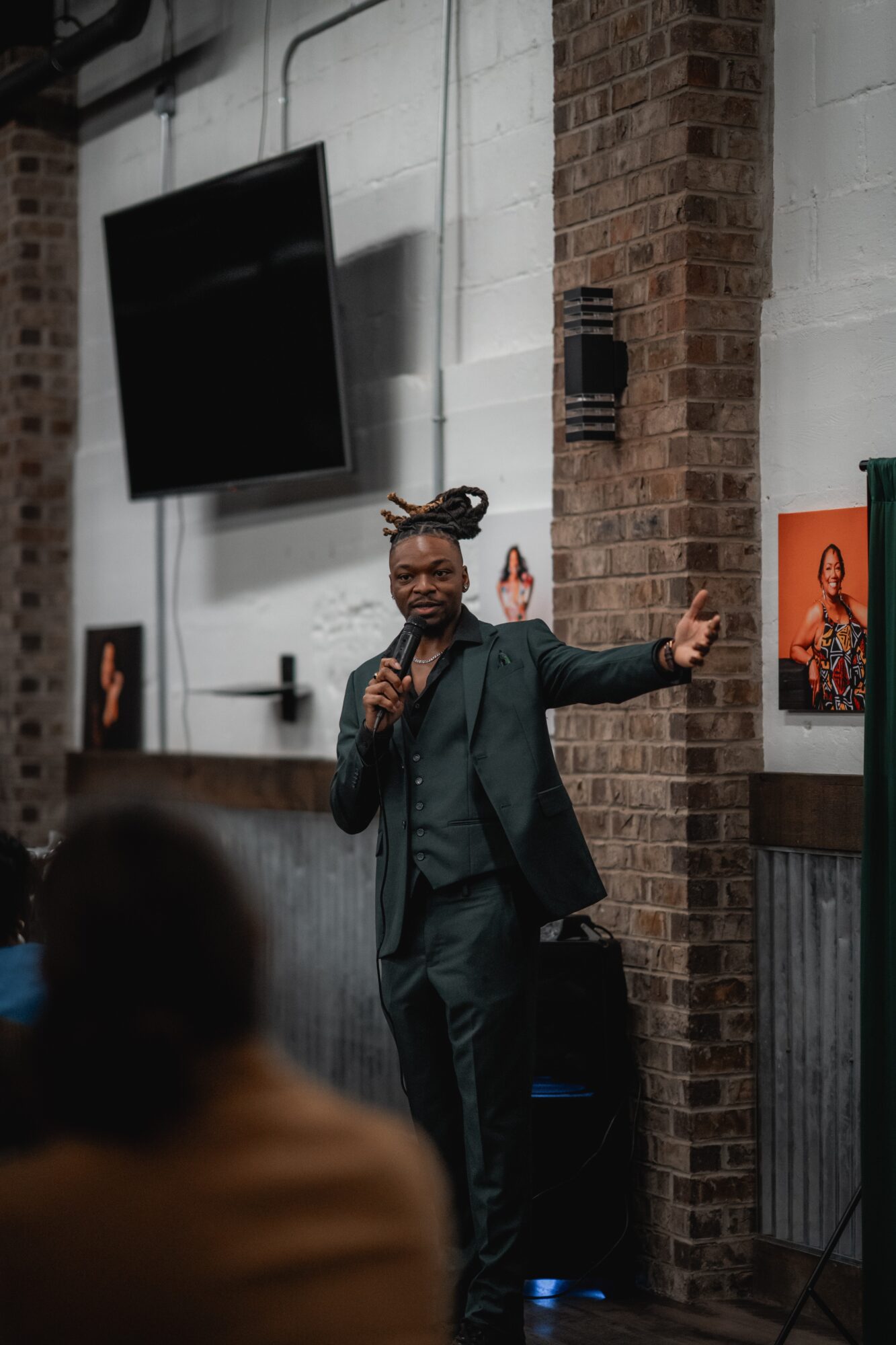Person with braided hair and glasses speaking into a microphone, gesturing with one hand, in a room with brick walls and artwork.