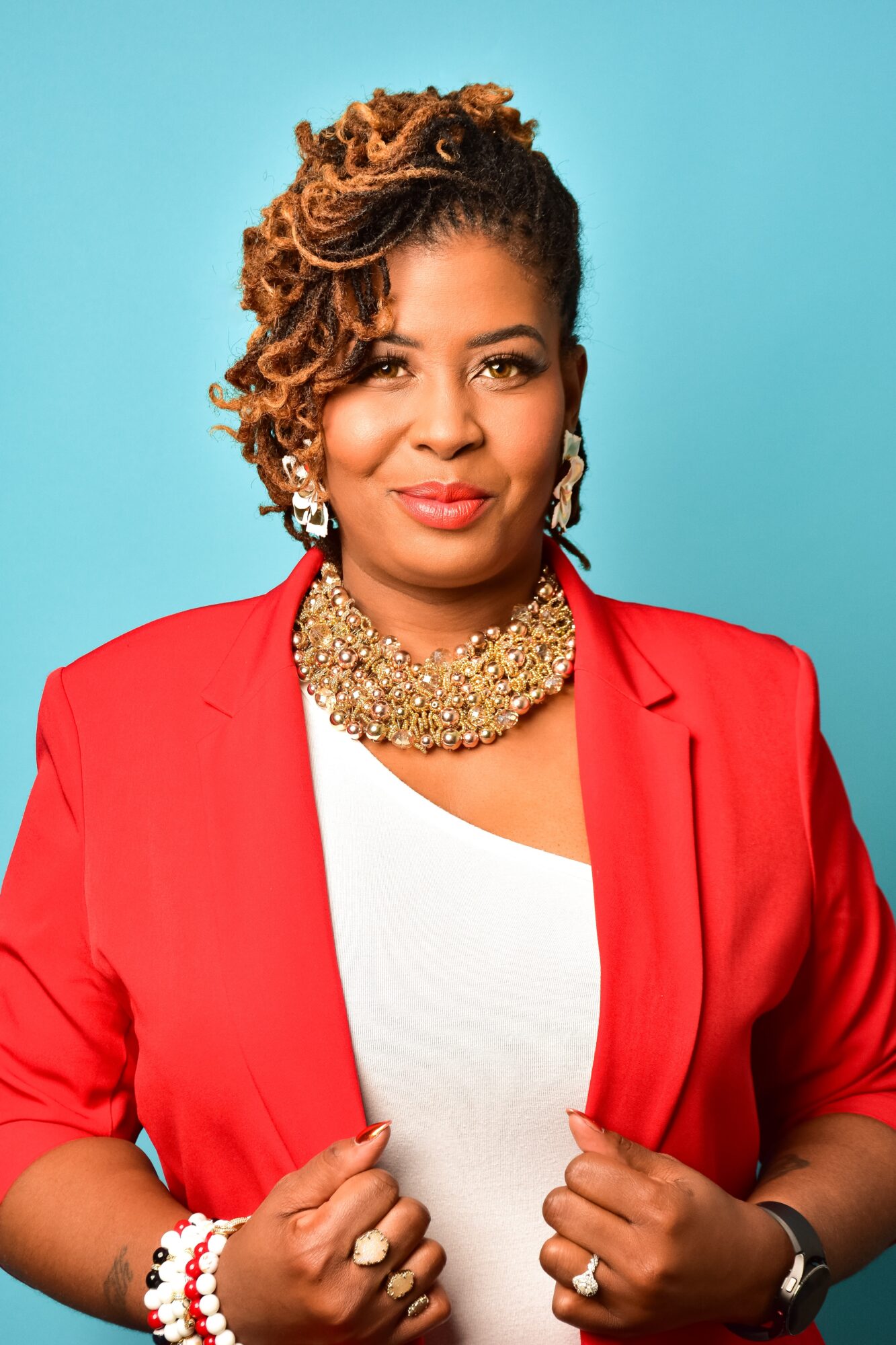 Woman with styled hair, wearing a red blazer, white top, and gold necklace, against a blue background.