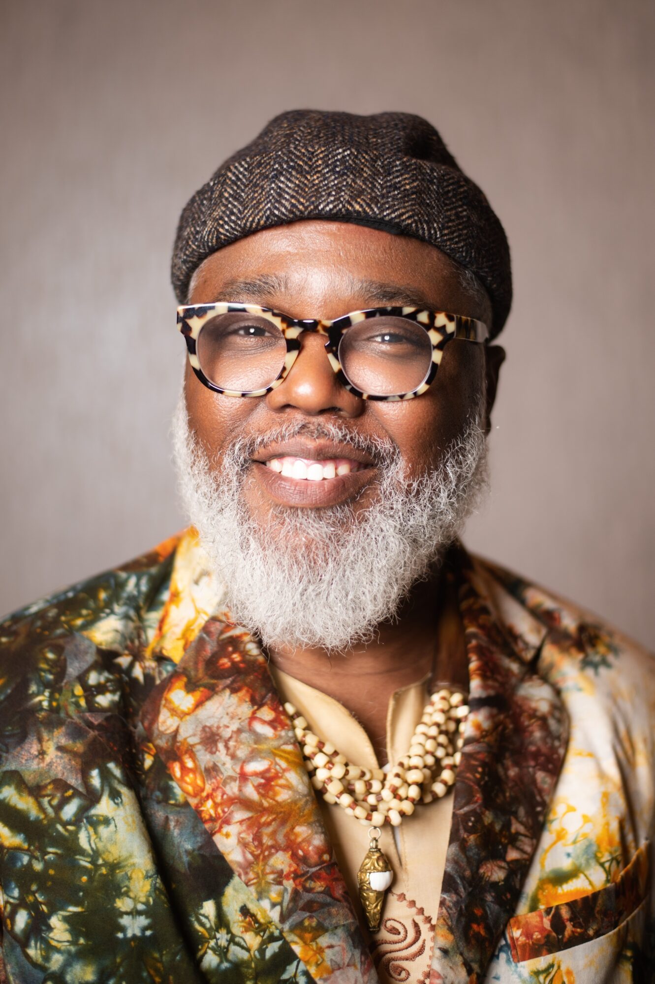 Man with glasses, gray beard, wearing a hat, patterned shirt, and necklace, smiling at the camera.