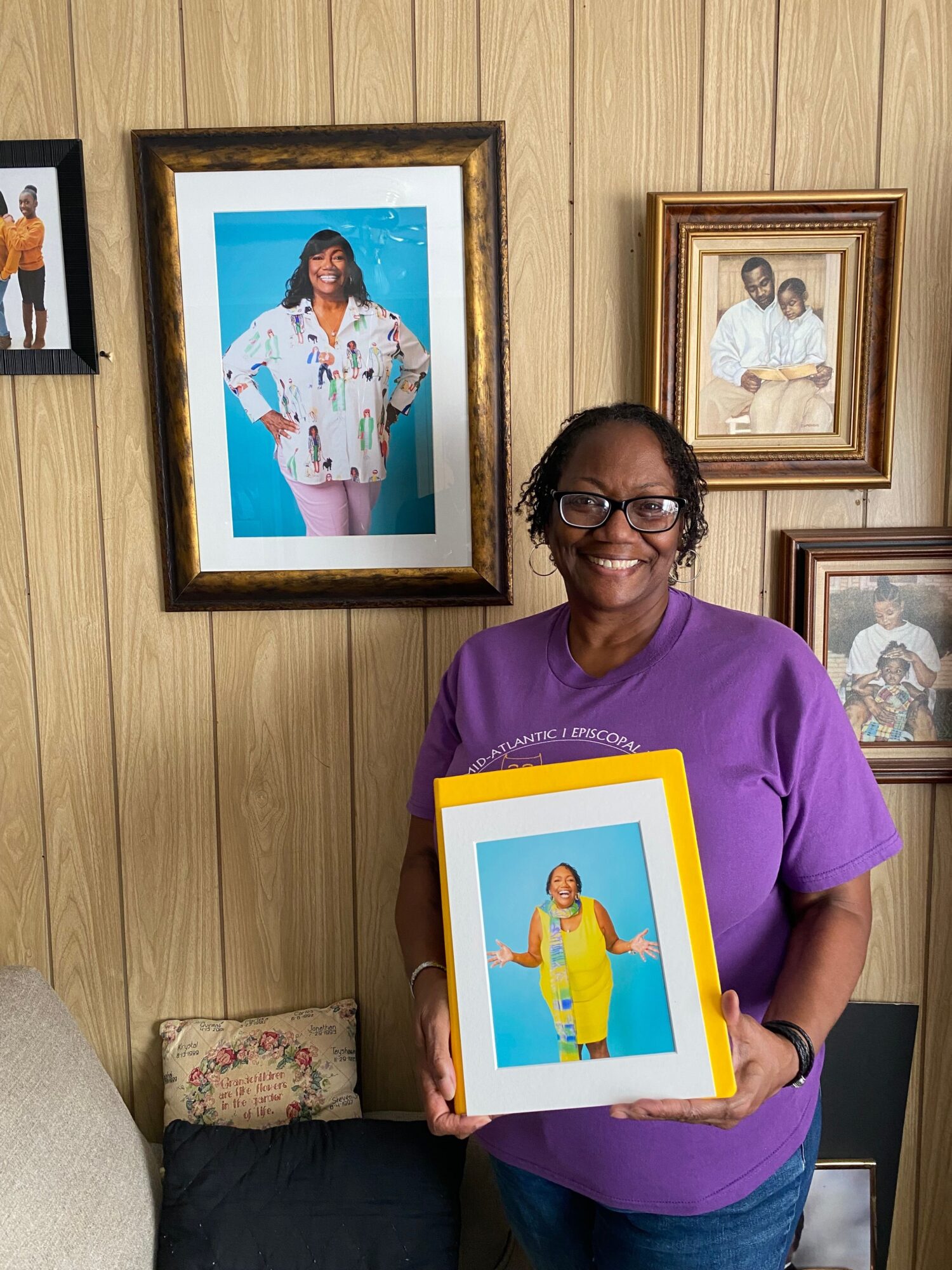 Woman holding a framed photo, standing next to framed pictures on a wooden wall, with a cushion nearby.