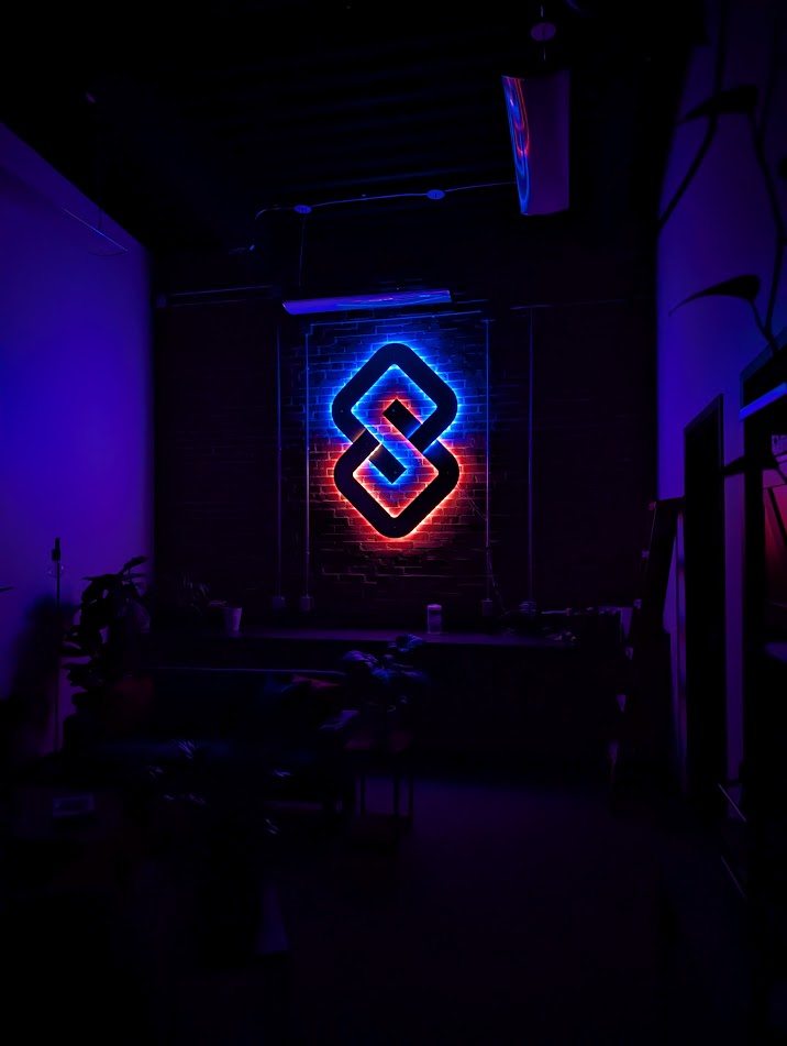 Neon sign with geometric design glowing in blue and red, mounted on a dark wall in a dimly lit room.