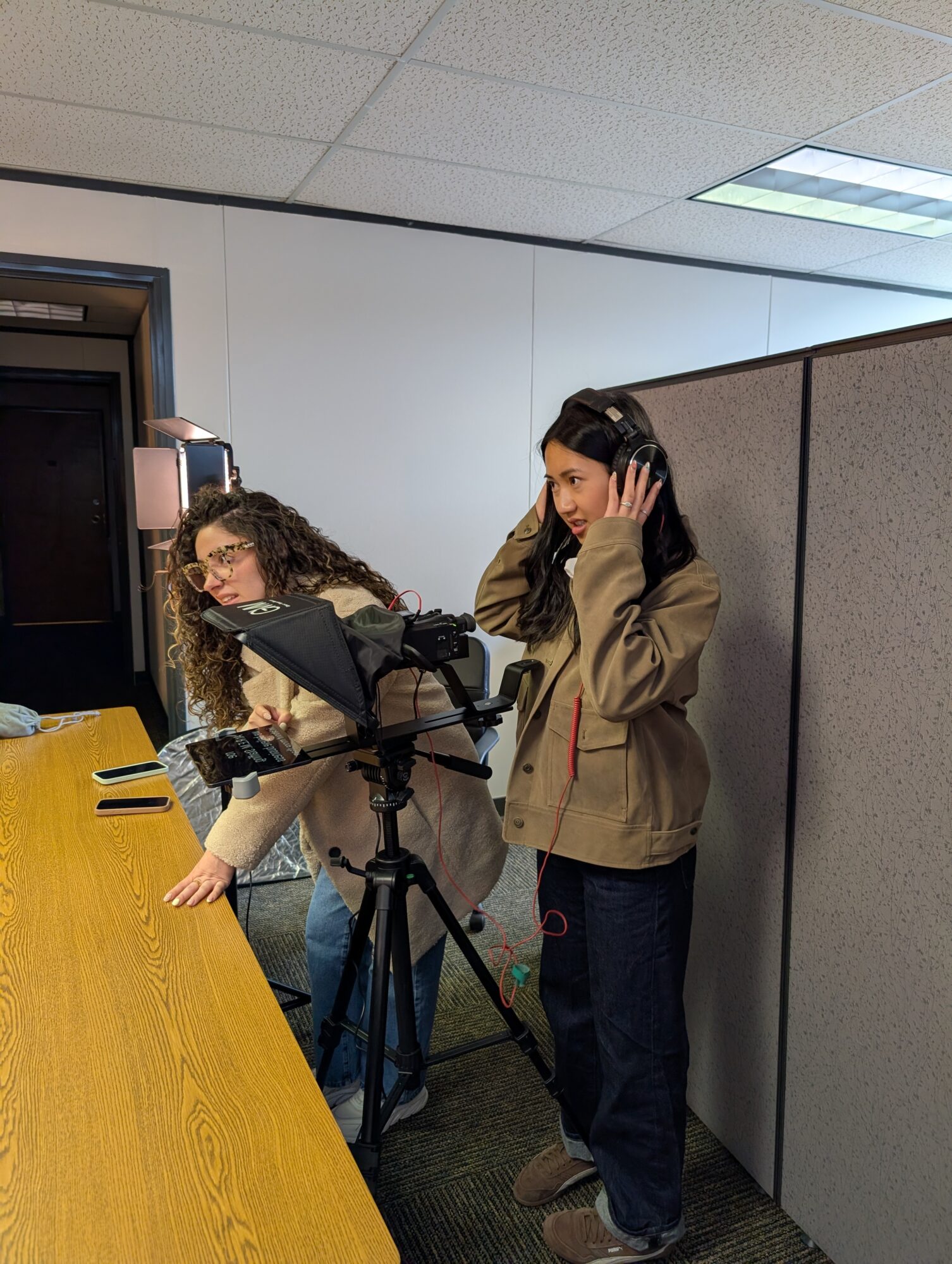 Two women working with audio equipment in an office, one adjusting a microphone and the other wearing headphones.