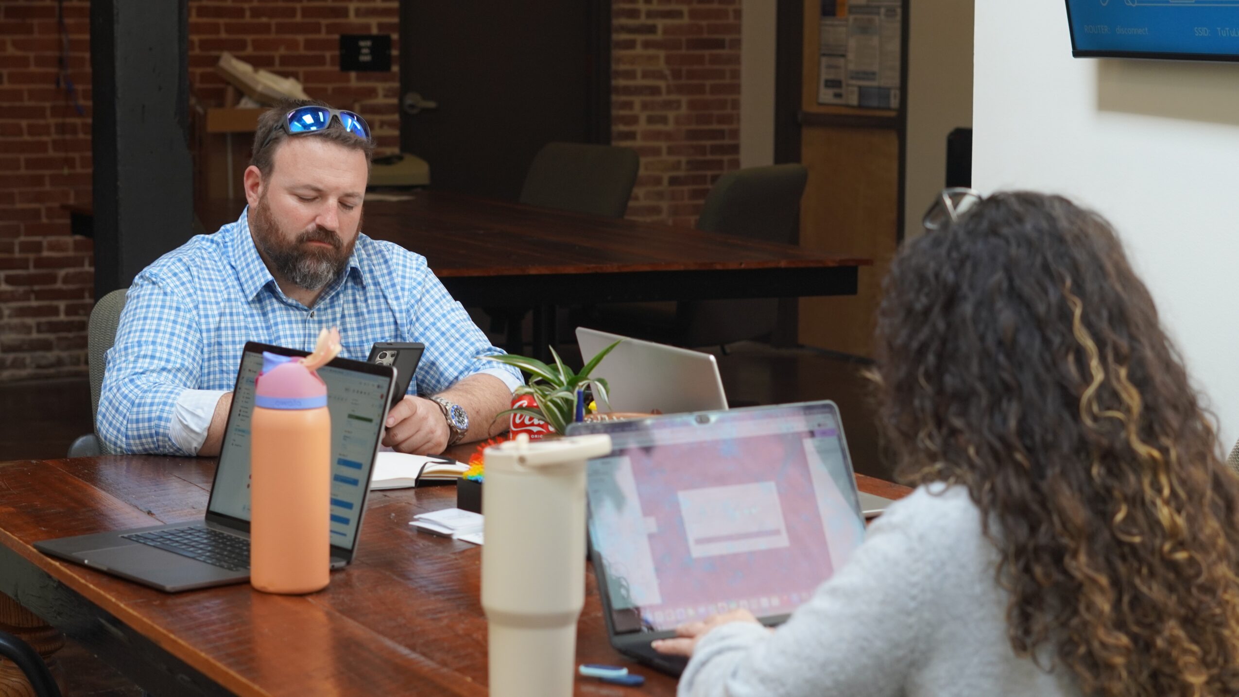 Two people working on laptops at a conference table in a room with brick walls and a TV screen.