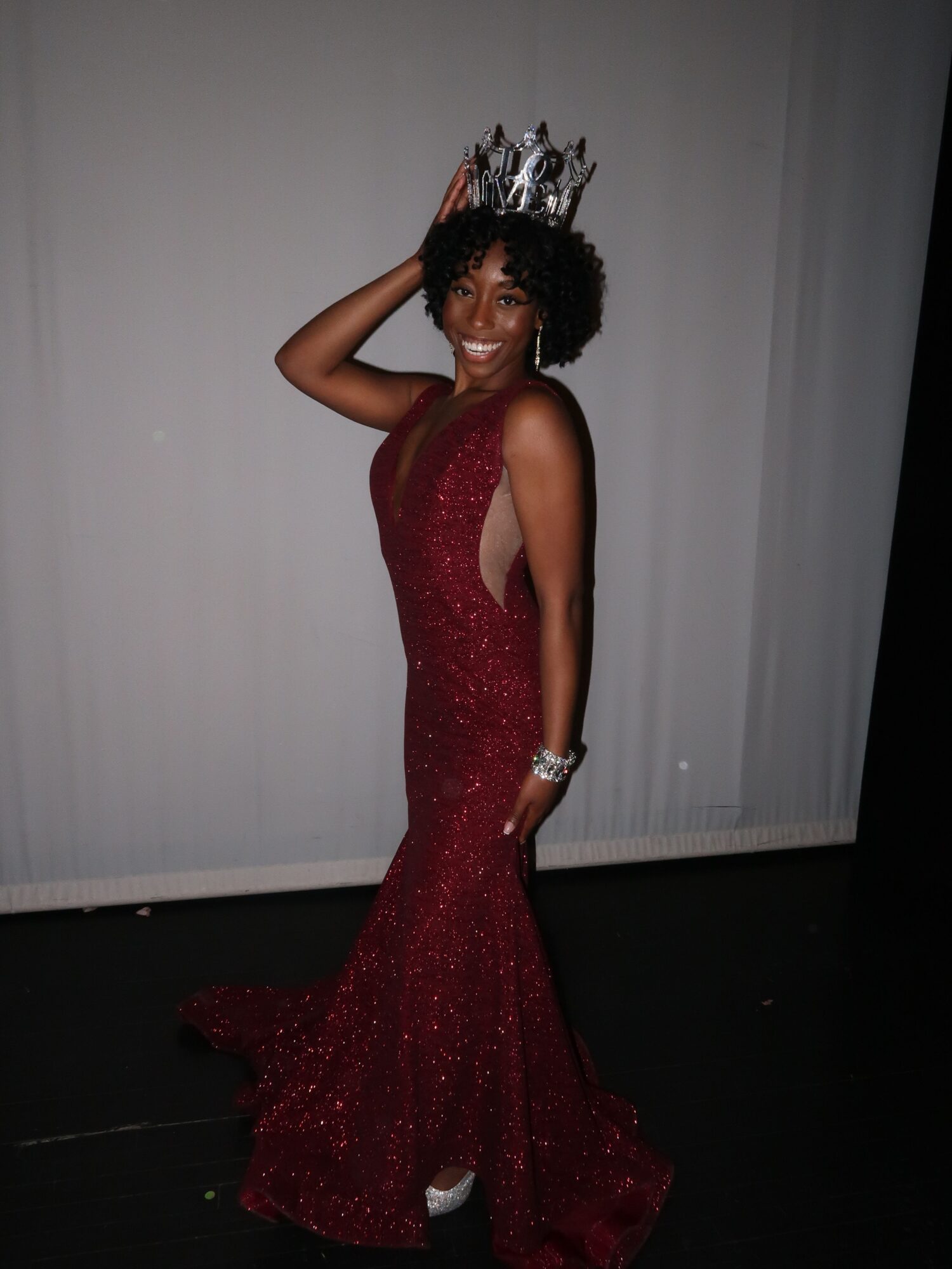Woman in a red gown with a crown, lying on the floor, posing with one arm behind her head, against a plain background.