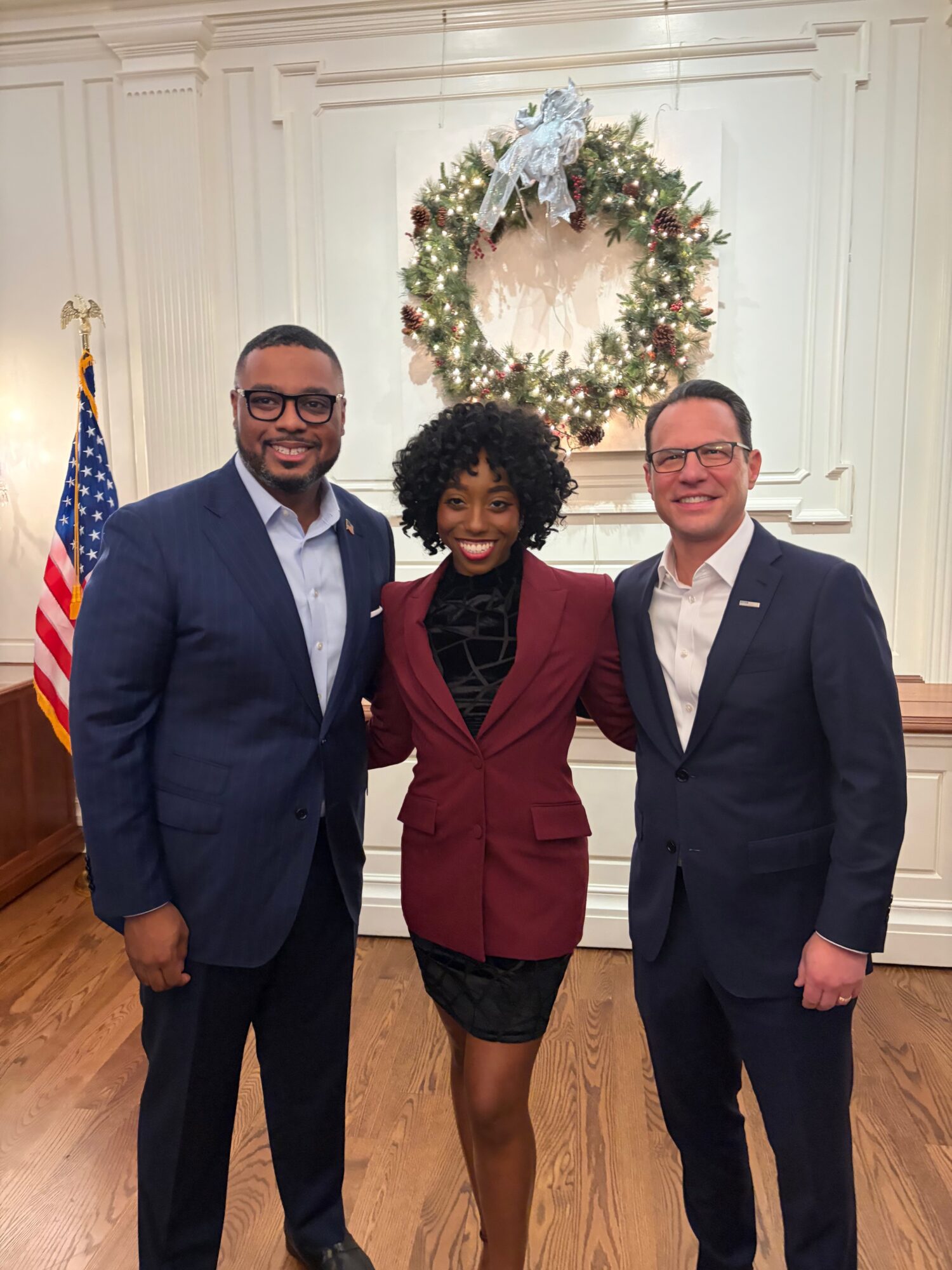 Three people in formal attire standing in front of a Christmas wreath and American flag, smiling.