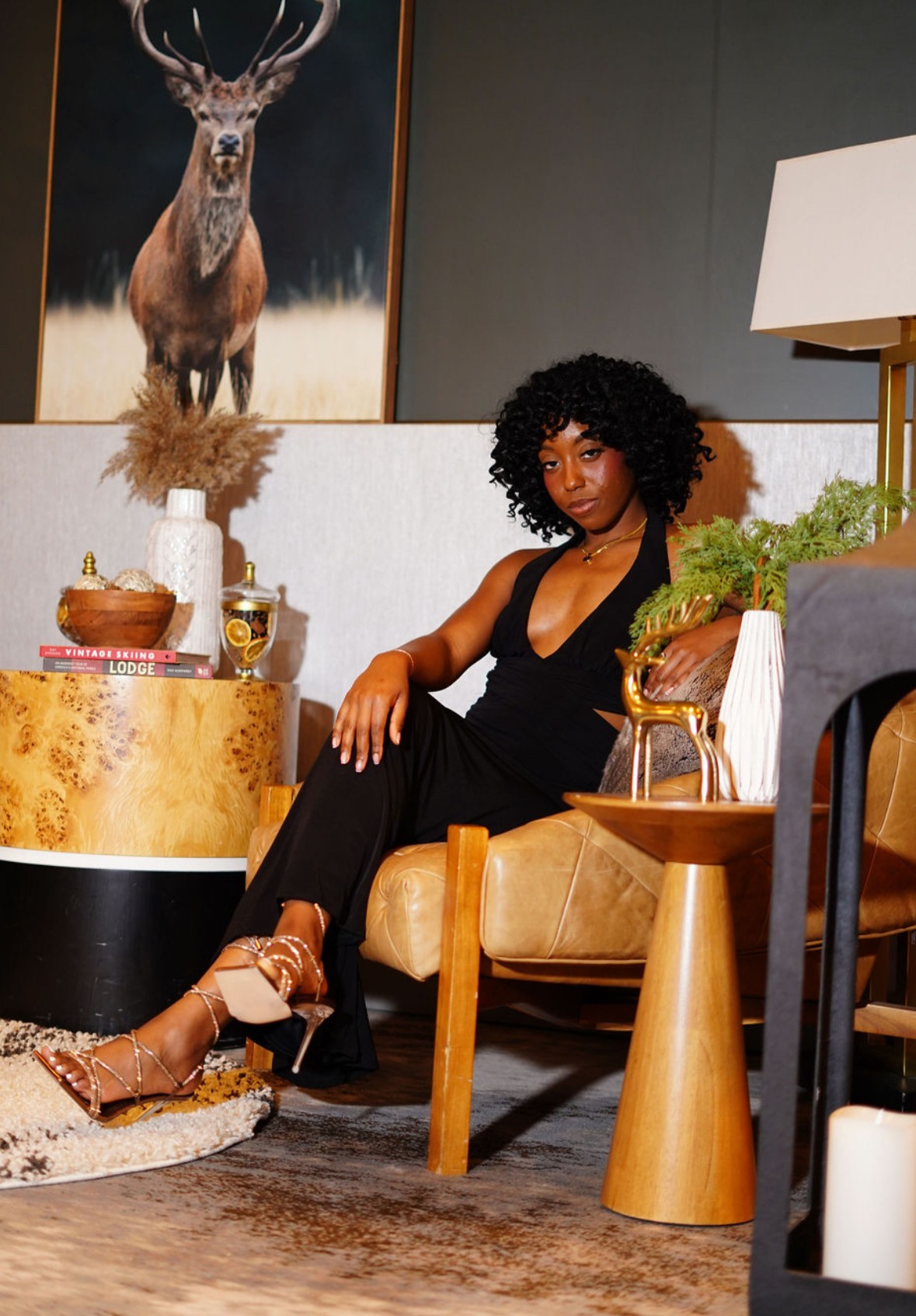 Woman with curly hair sitting on a tan chair in a room with artwork, plants, and furniture, wearing a black outfit.