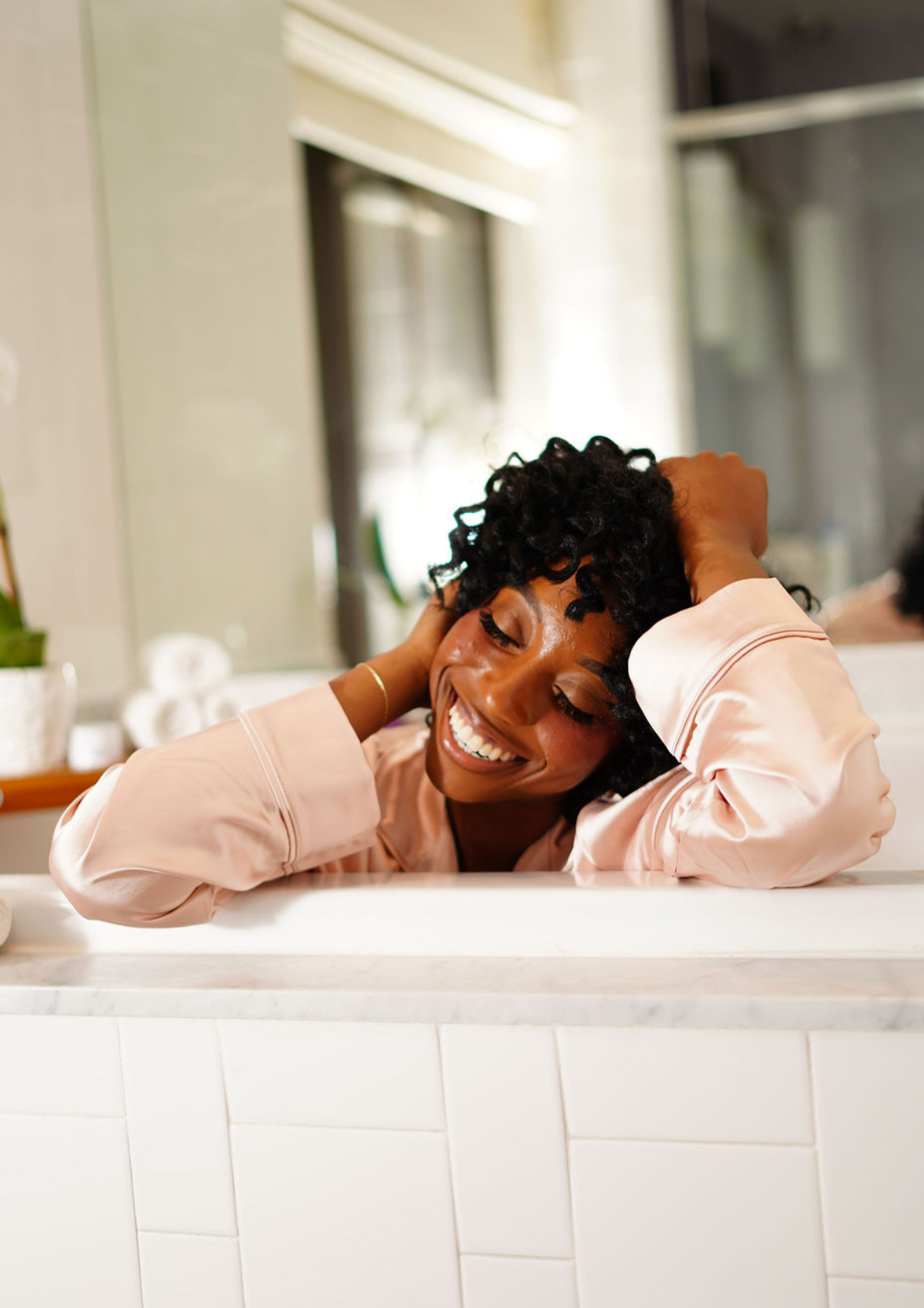 Smiling woman with curly hair resting her head on her arms at a kitchen counter.