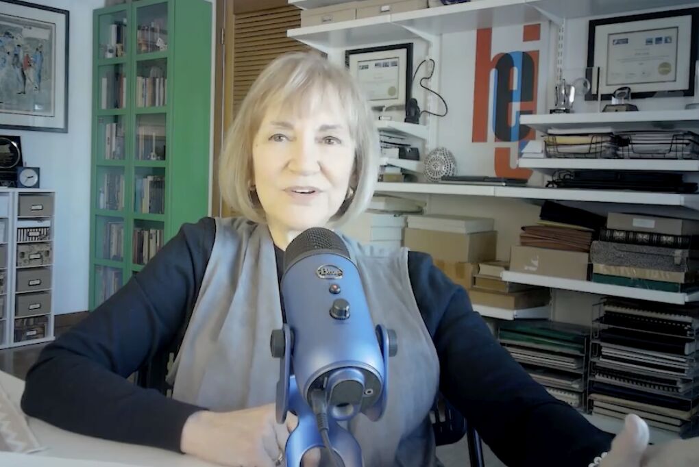 Woman speaking into a microphone in a room with shelves and framed certificates behind her.