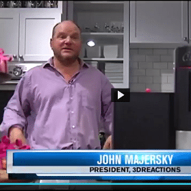 Man in purple shirt standing in a kitchen with cabinets and appliances, speaking on a video segment, with a news banner.