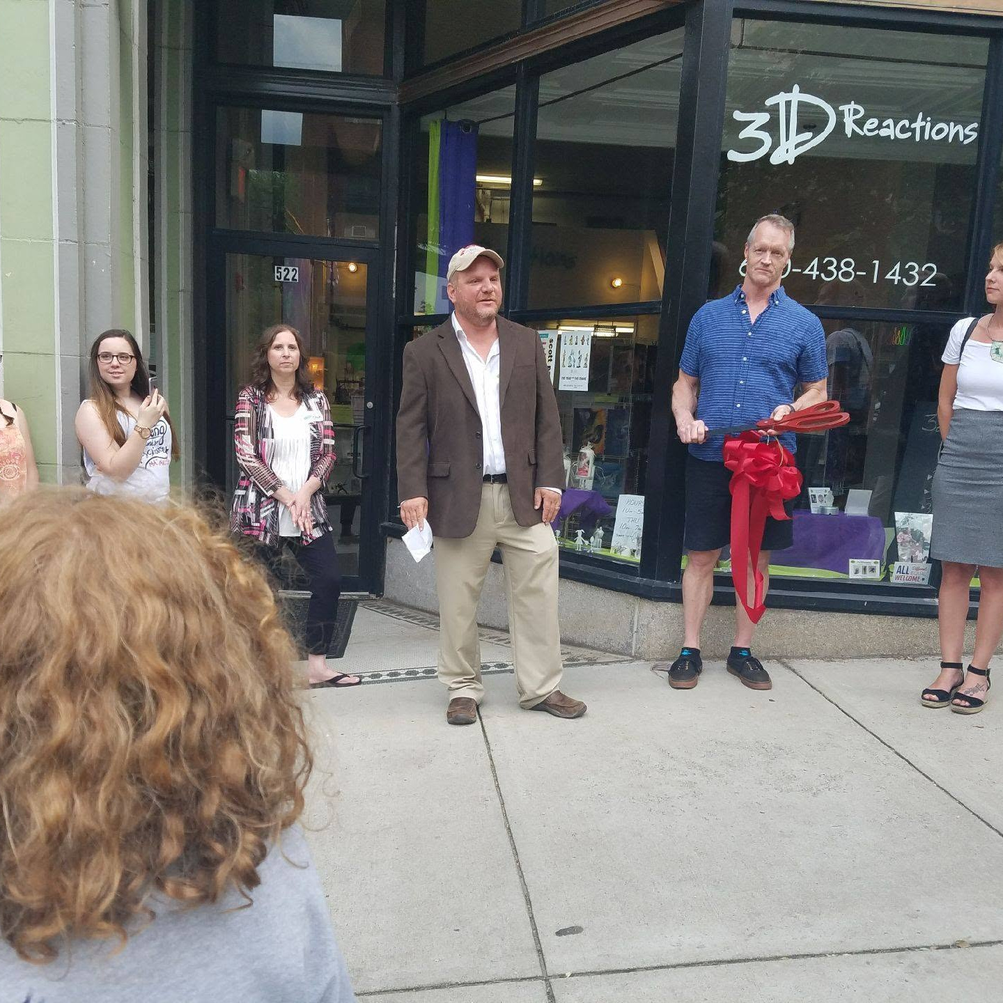 Group of people standing outside a storefront with glass windows, some holding items, in daytime.